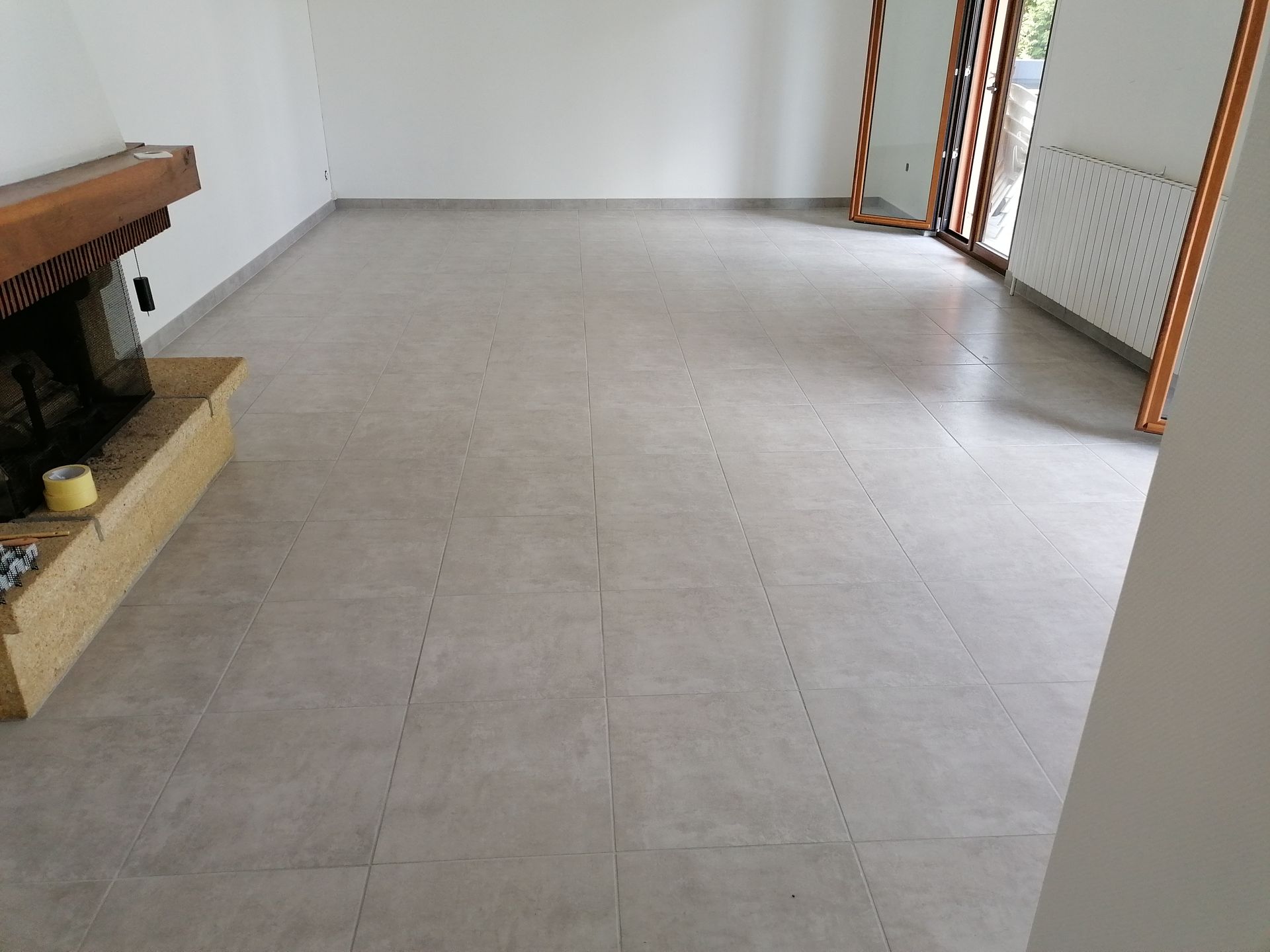 Un salon avec un carrelage carré gris clair, une cheminée à gauche et une porte-fenêtre coulissante à droite.