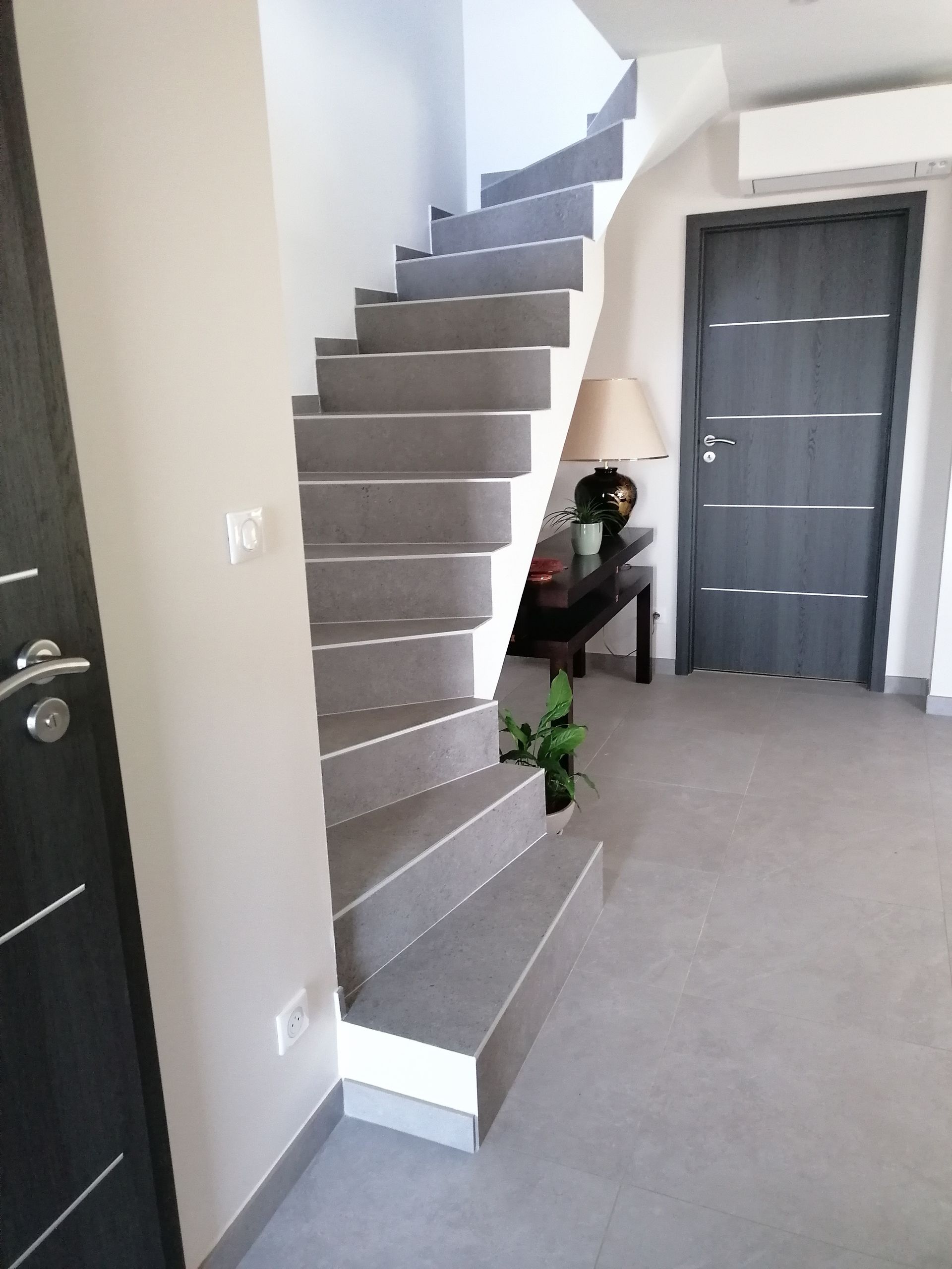 Un escalier aux marches grises et contremarches blanches se trouve à côté d'une porte sombre et d'une petite table d'appoint avec une lampe.