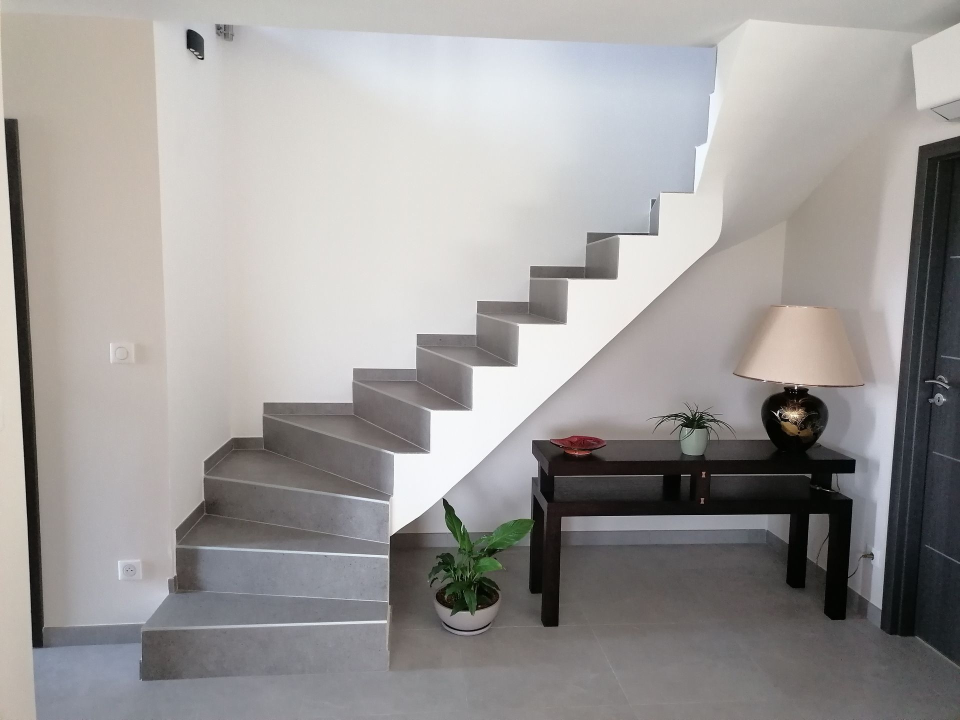 Un escalier aux marches grises et aux murs blancs mène à l'étage. Une table sombre et une lampe se trouvent à proximité.