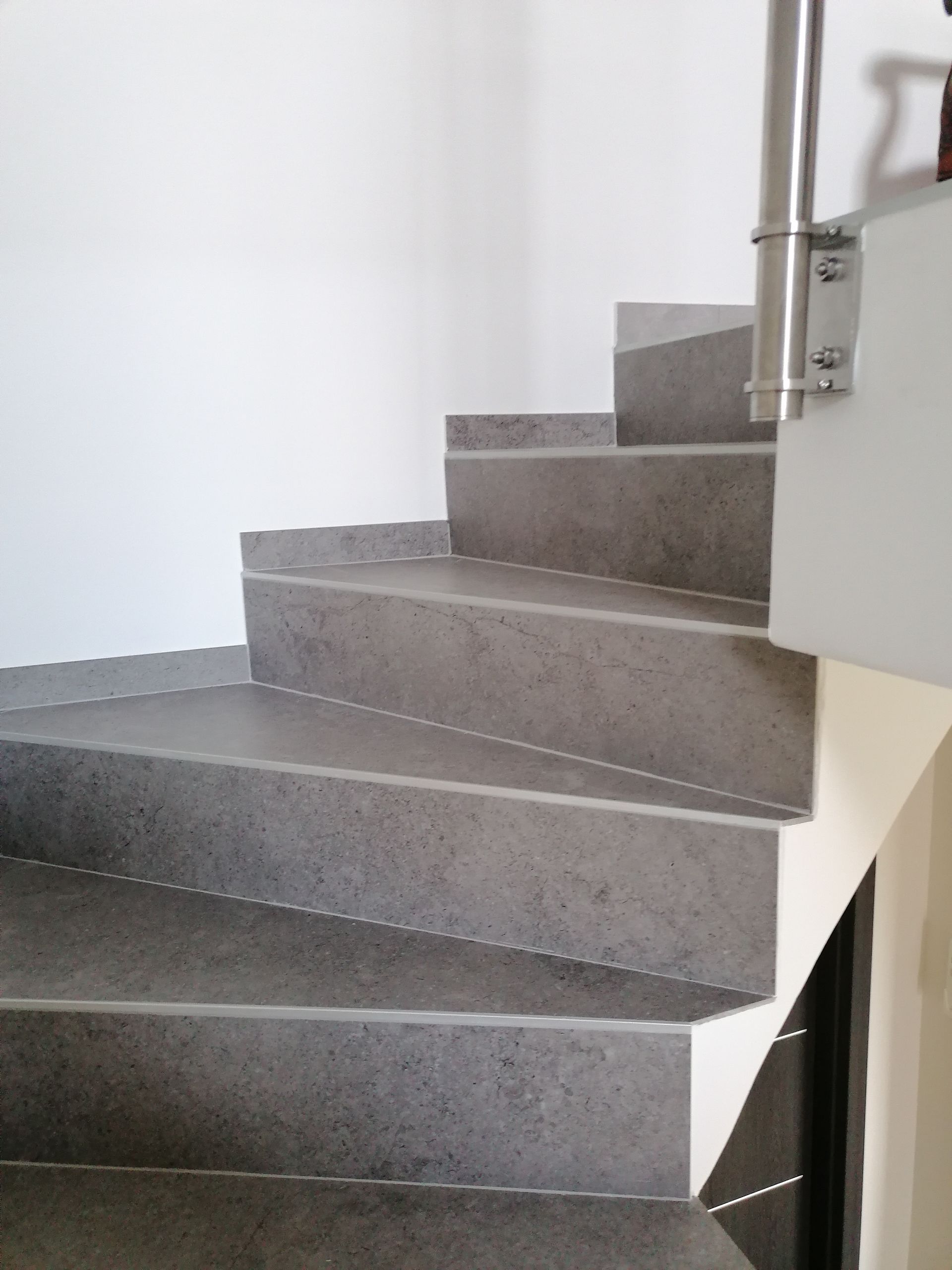 Un escalier en colimaçon aux marches carrelées de gris et à la rampe en métal argenté fixée contre un mur blanc.