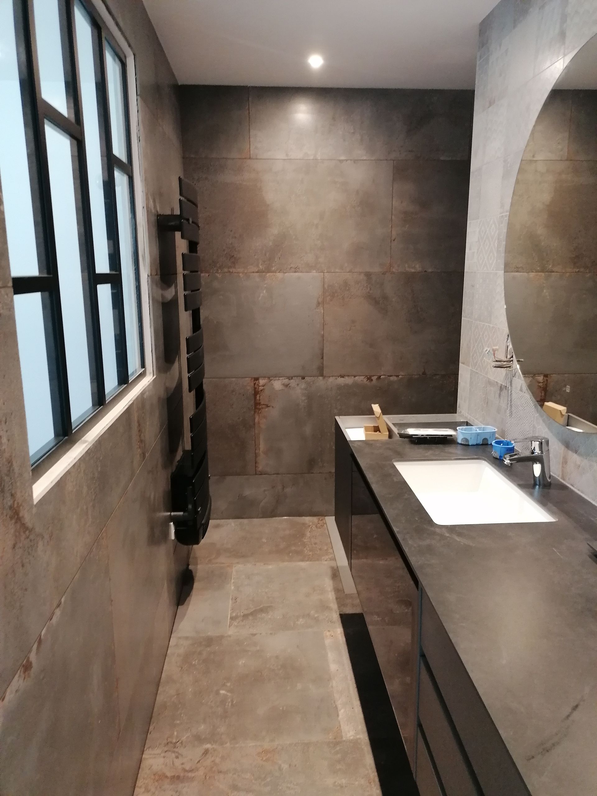 Salle de bains moderne aux murs gris foncé, long comptoir avec lavabo et miroir rond. Fenêtre à gauche.