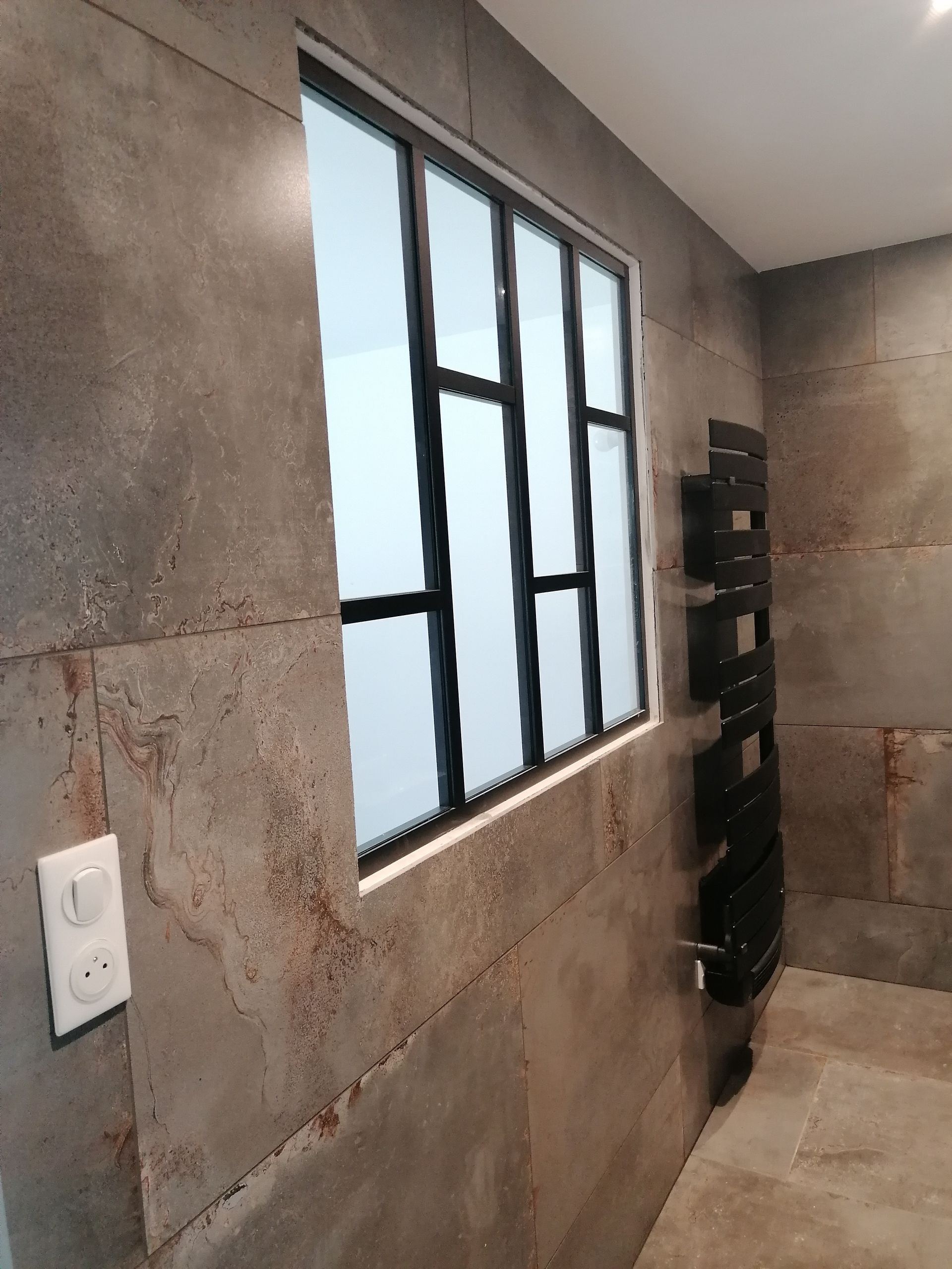 Une fenêtre à cadre métallique noir encastrée dans un mur recouvert de carreaux bruns à l'aspect vieilli, dans une pièce.