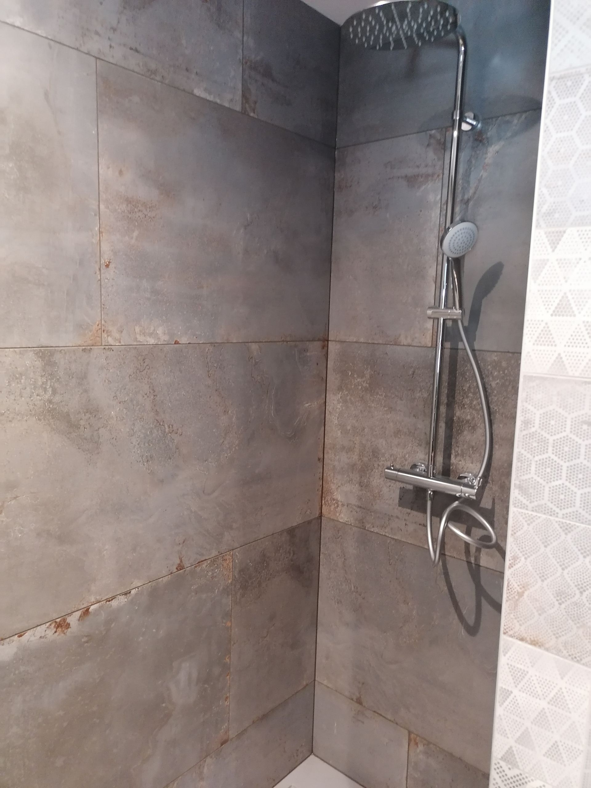 Une cabine de douche moderne avec de grands carreaux rectangulaires gris imitation pierre et une pomme de douche chromée effet pluie.