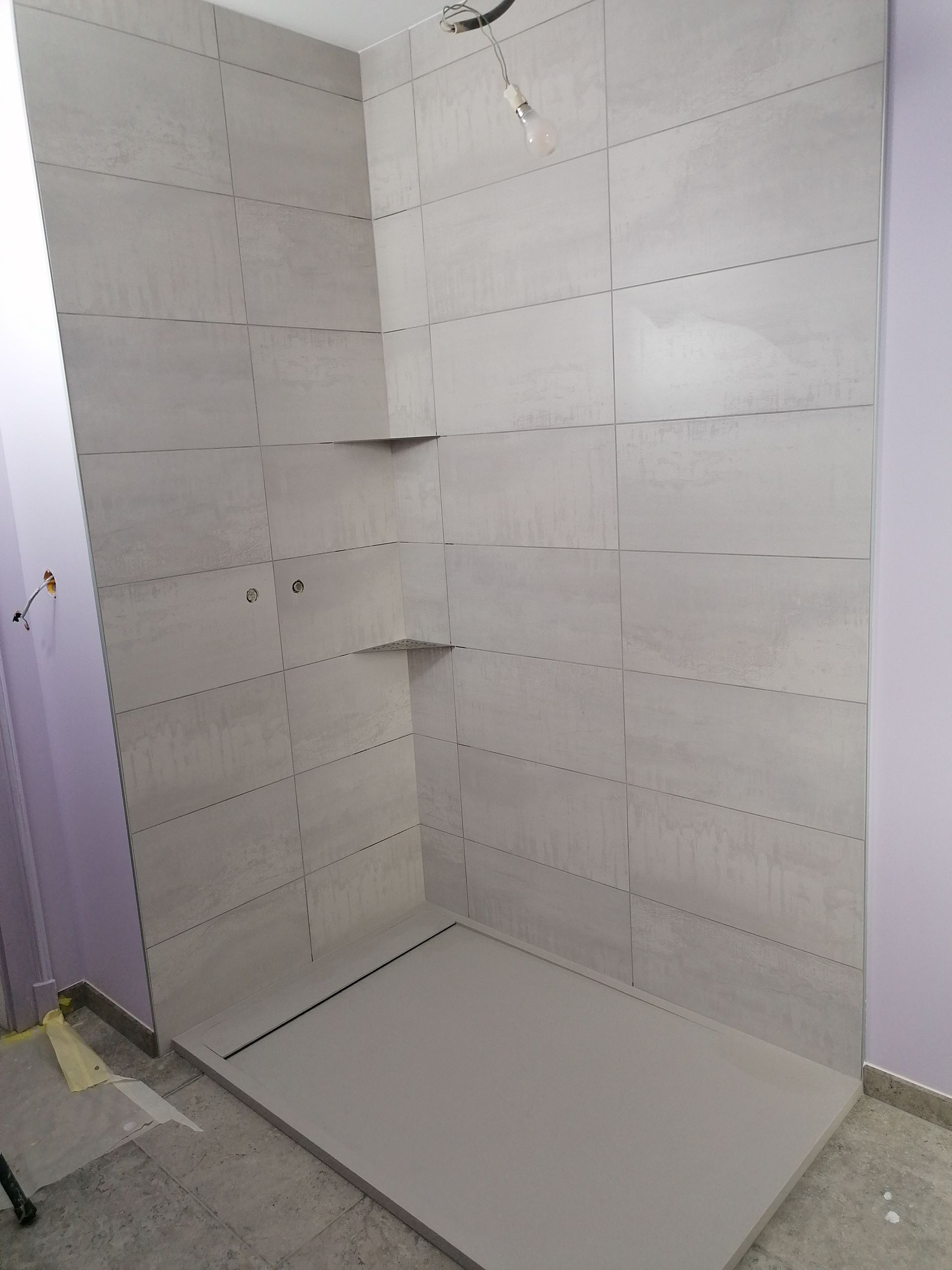 Une cabine de douche d'angle en construction avec des murs carrelés gris clair, deux étagères d'angle intégrées et un receveur de douche gris.