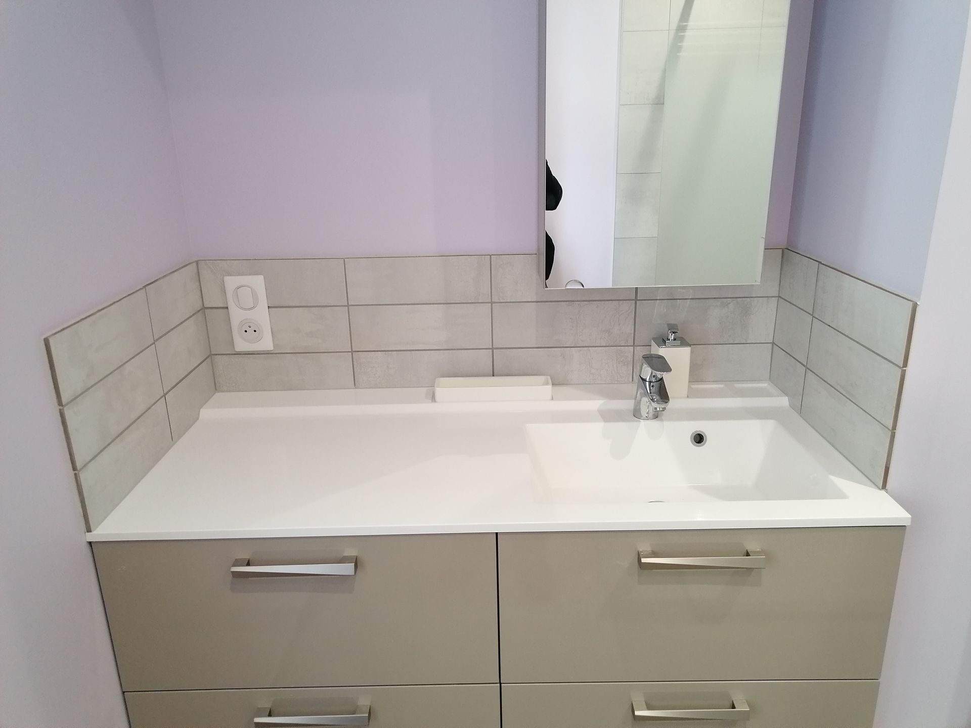 Meuble-lavabo de salle de bain avec un comptoir blanc, un lavabo rectangulaire, des armoires gris clair et une crédence en carrelage gris avec une prise électrique.
