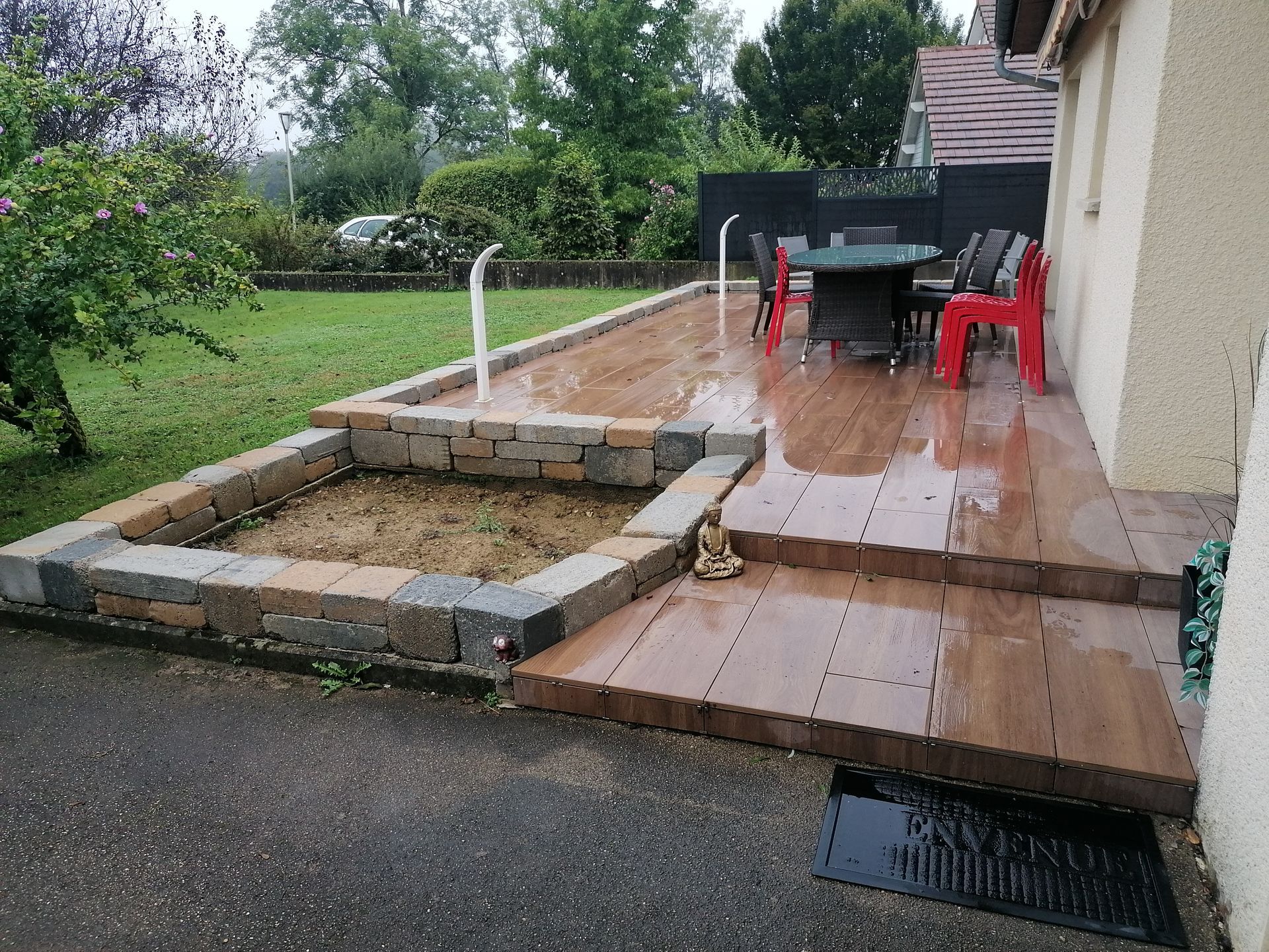 Une terrasse en bois avec une jardinière en pierre, un ensemble de salle à manger avec des chaises rouges et une petite statue de Bouddha devant une maison.