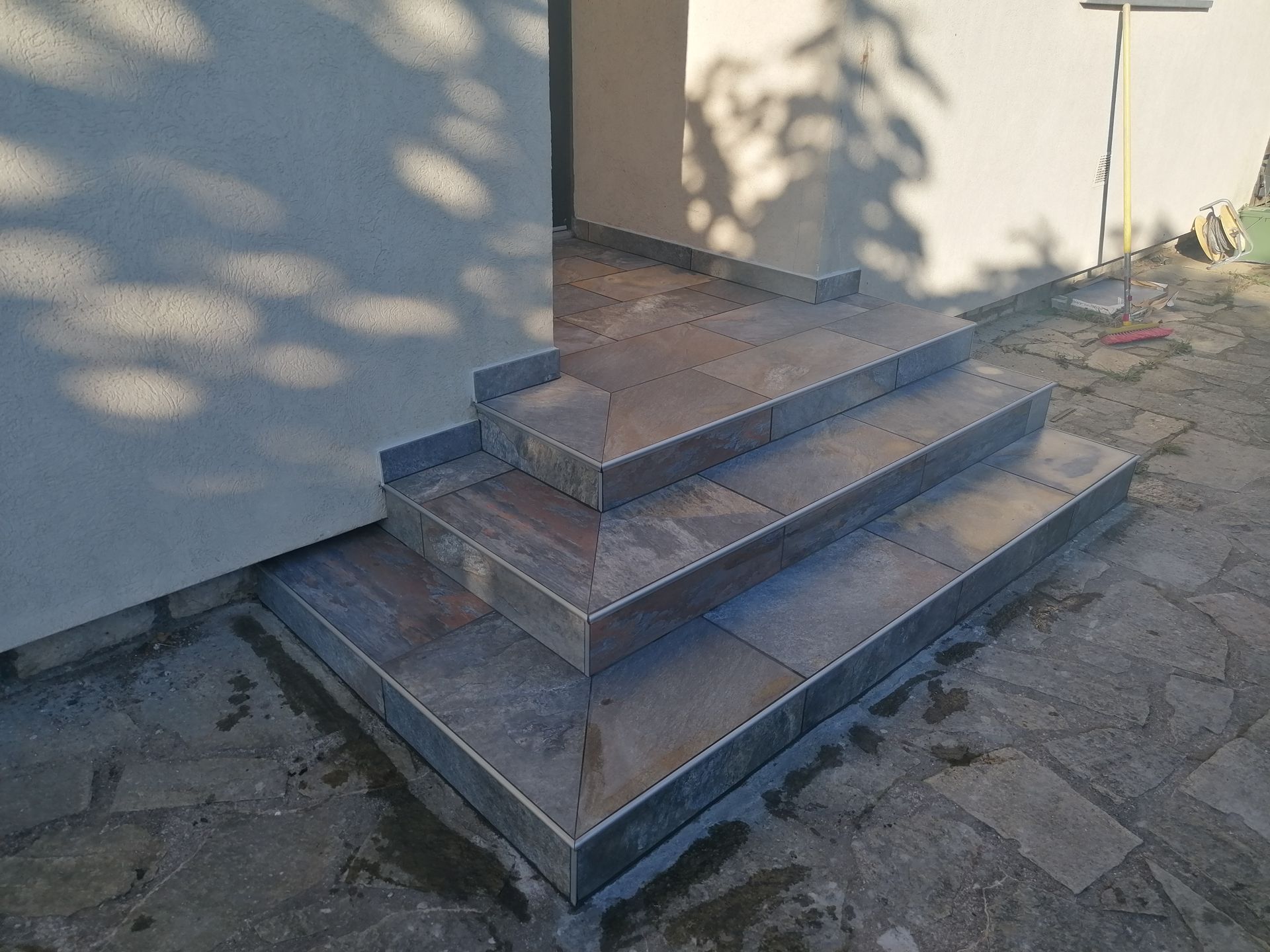 Trois marches carrelées de pierre brunâtre et bordées de métal mènent à une porte, adossée à un mur extérieur de couleur claire.