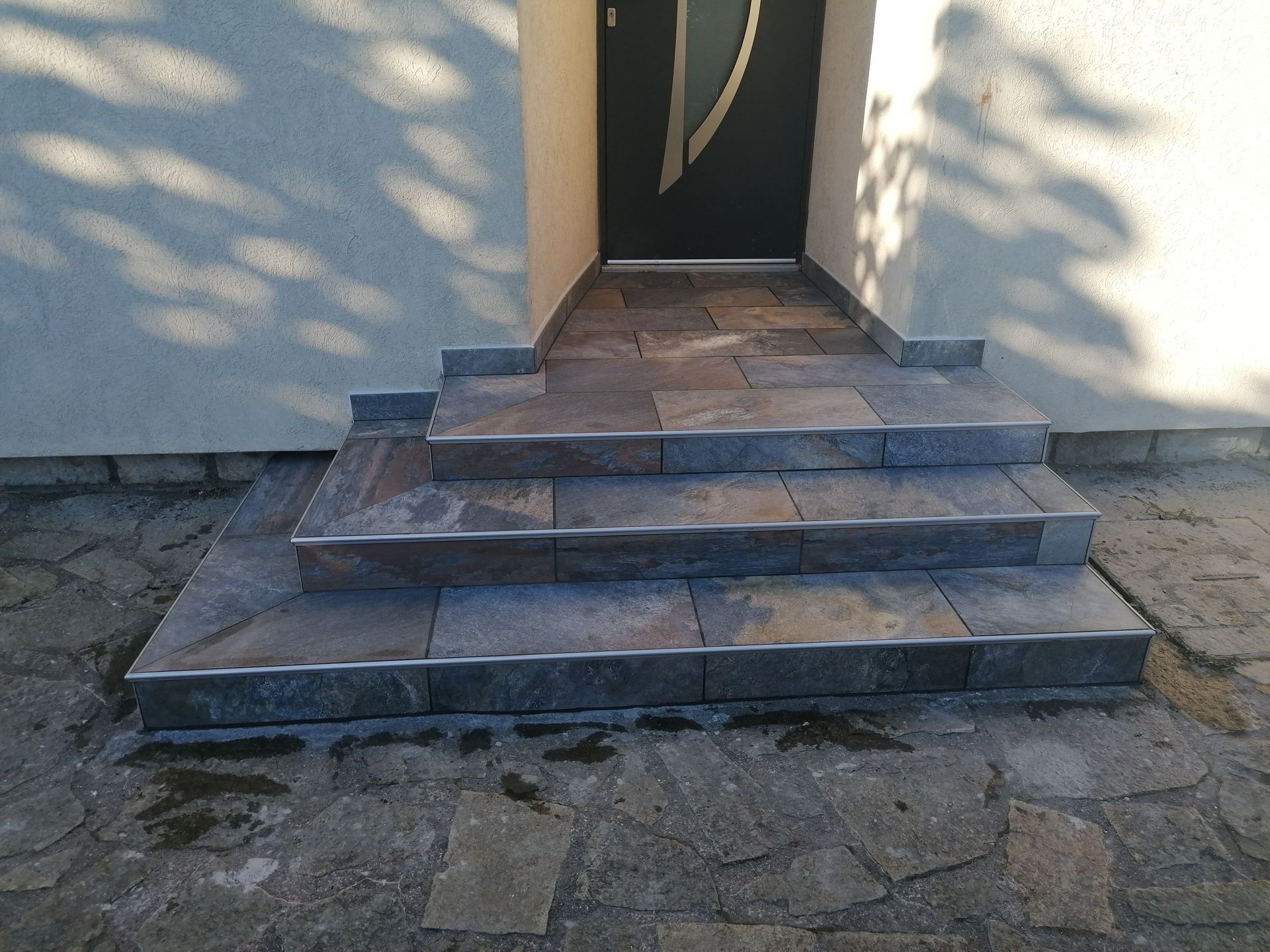 Un escalier de trois marches carrelées menant à une porte d'entrée sombre sur la façade claire d'un bâtiment.