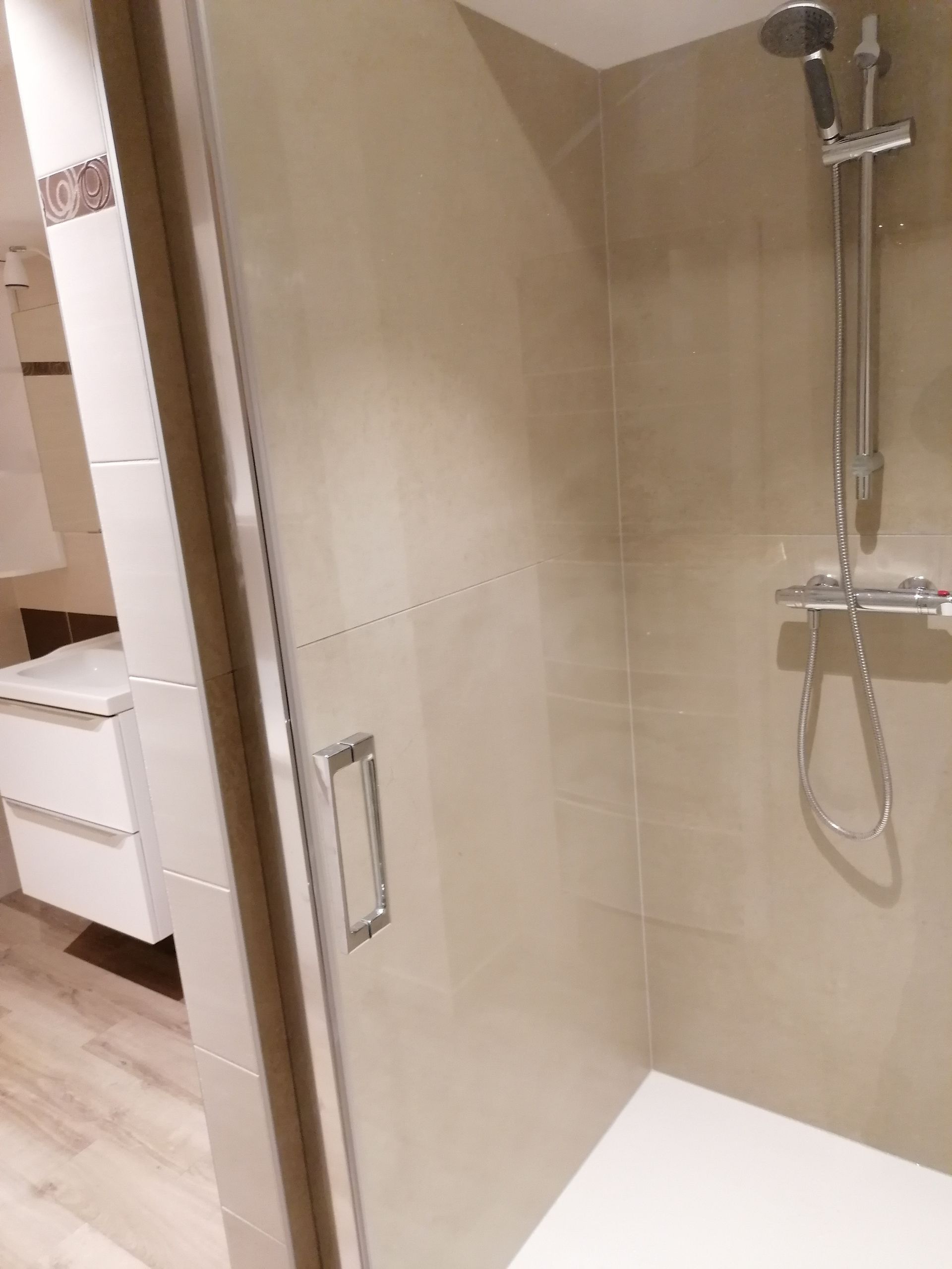 Une cabine de douche moderne avec porte en verre, pommeau de douche chromé et murs carrelés beiges dans une salle de bains lumineuse et contemporaine.