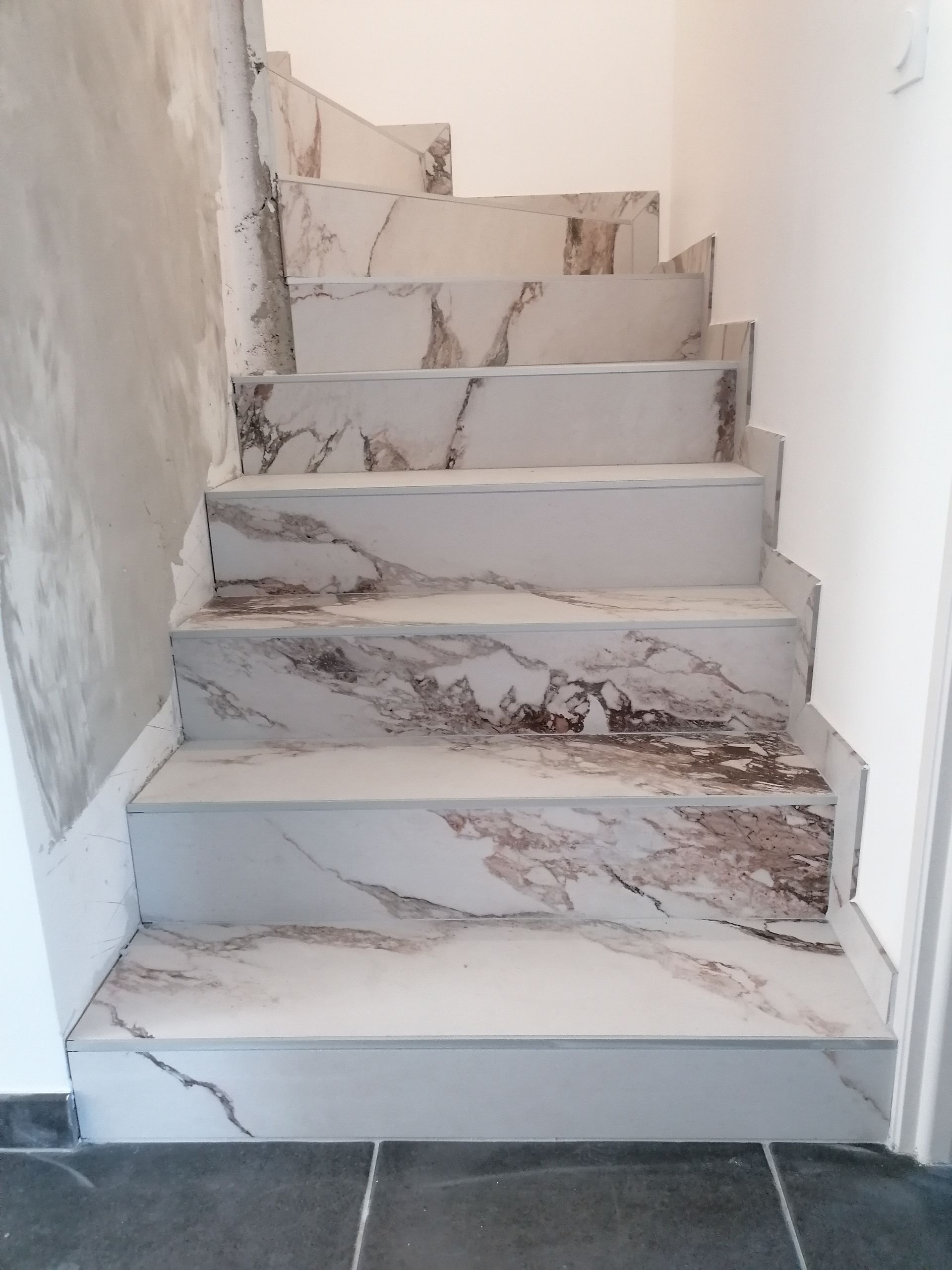 Escalier carrelé en marbre sur fond de murs blancs. Un sol gris foncé mène aux marches. Certaines marches présentent des traces de brunissement.