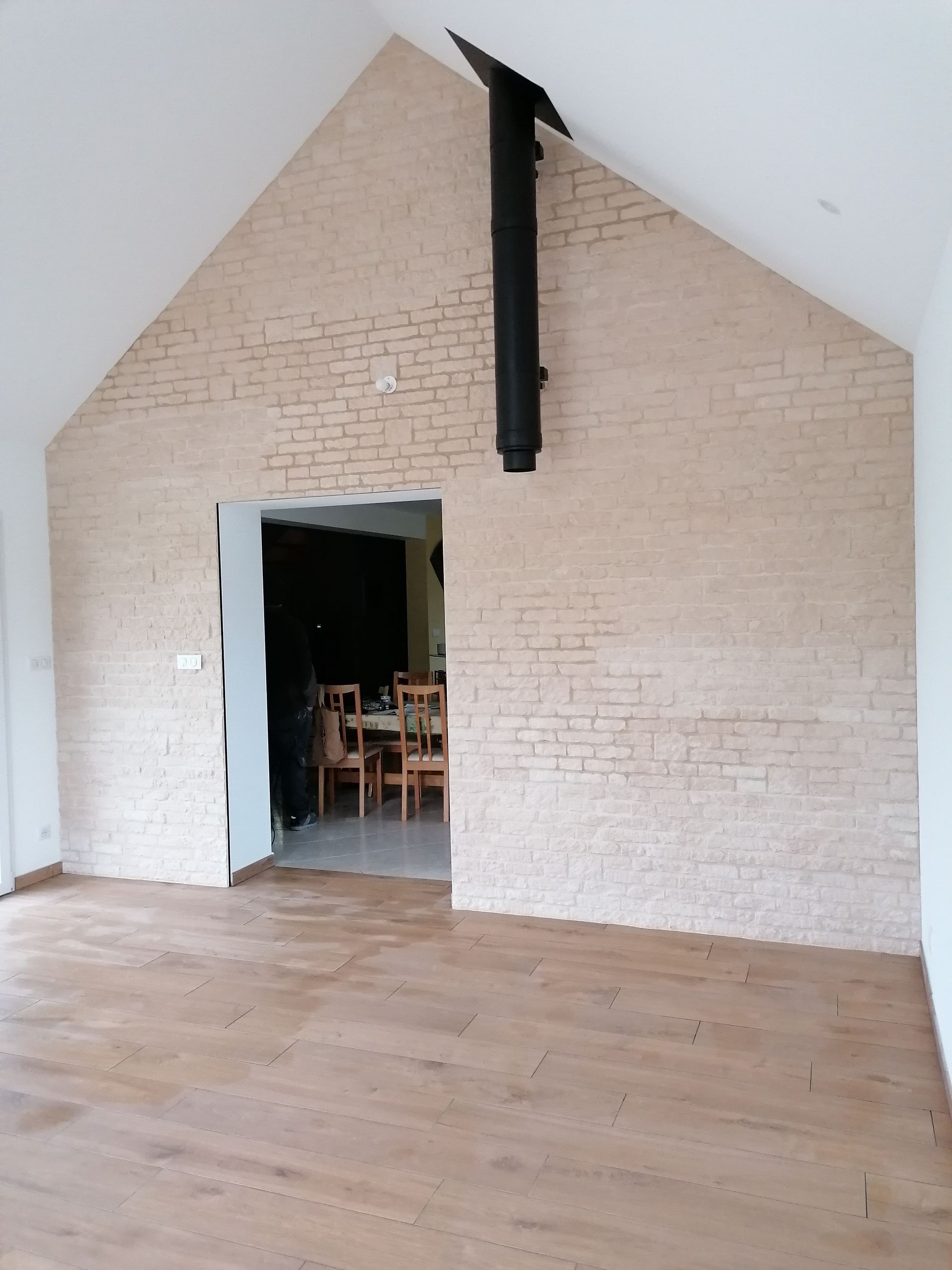 Intérieur d'une pièce avec parquet clair, mur de briques, porte, cheminée noire et plafond voûté.