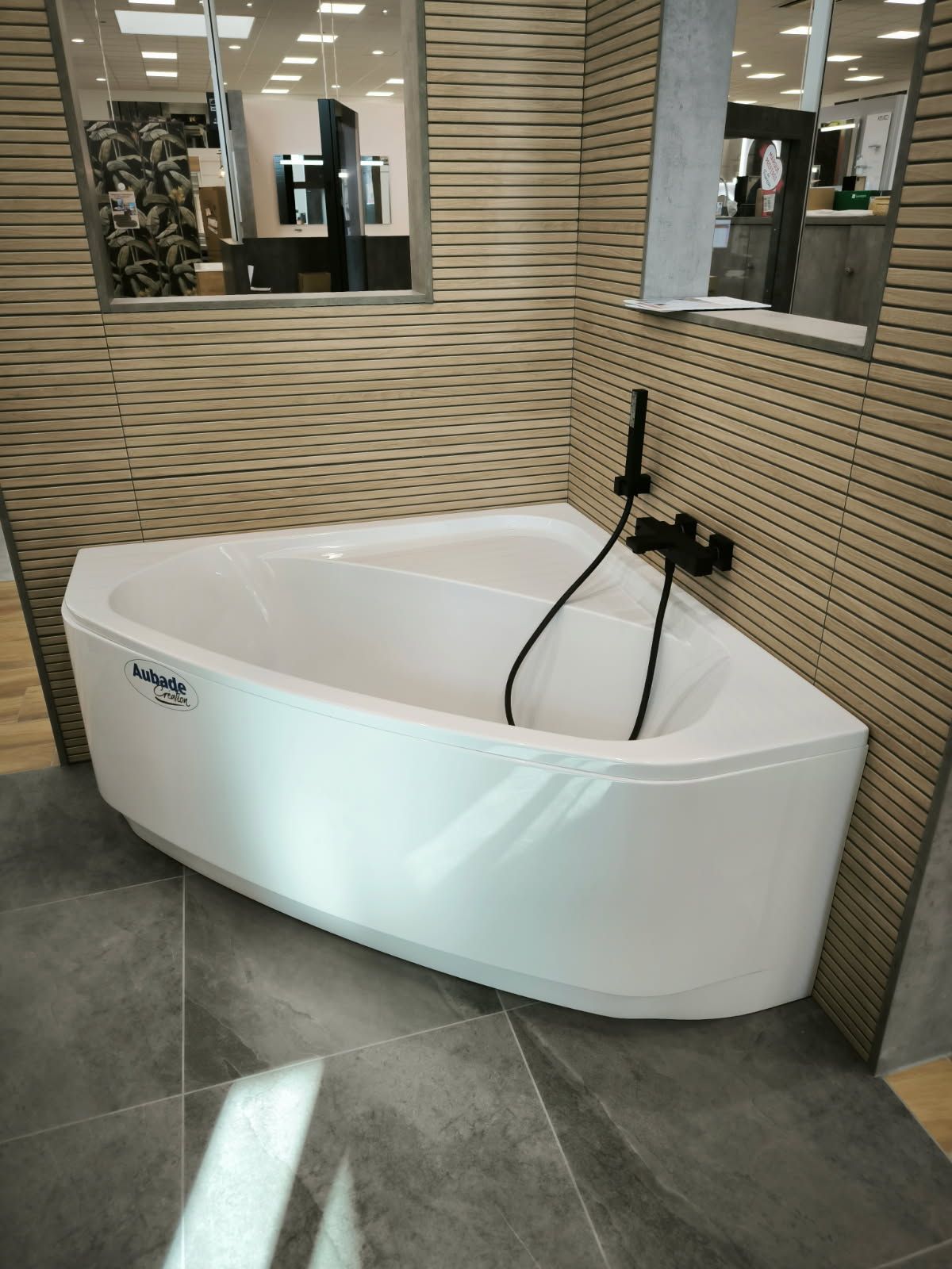 Dans une salle d'exposition, une baignoire d'angle blanche avec robinetterie noire se détache sur un mur carrelé de couleur beige ondulée et un sol carrelé de gris.