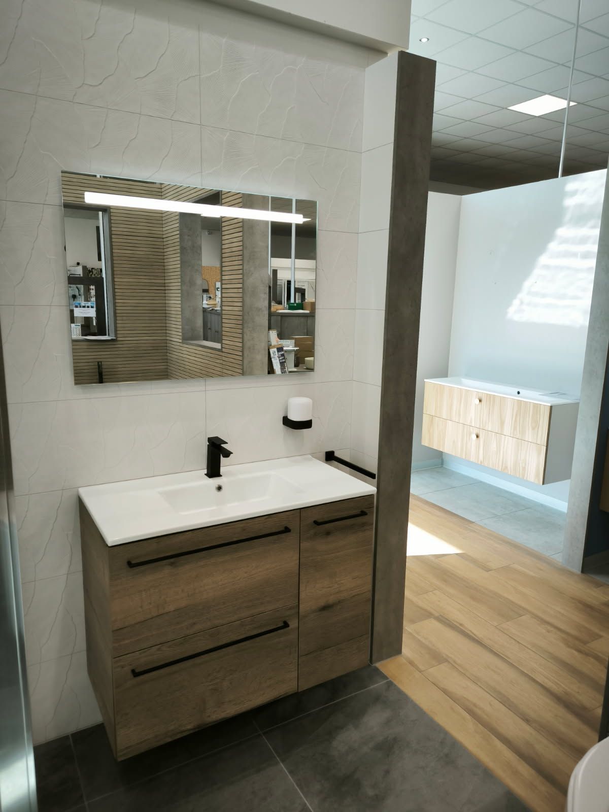 Meuble-lavabo moderne avec armoires en bois, lavabo blanc, robinet noir et miroir rectangulaire, exposé dans une salle d'exposition.