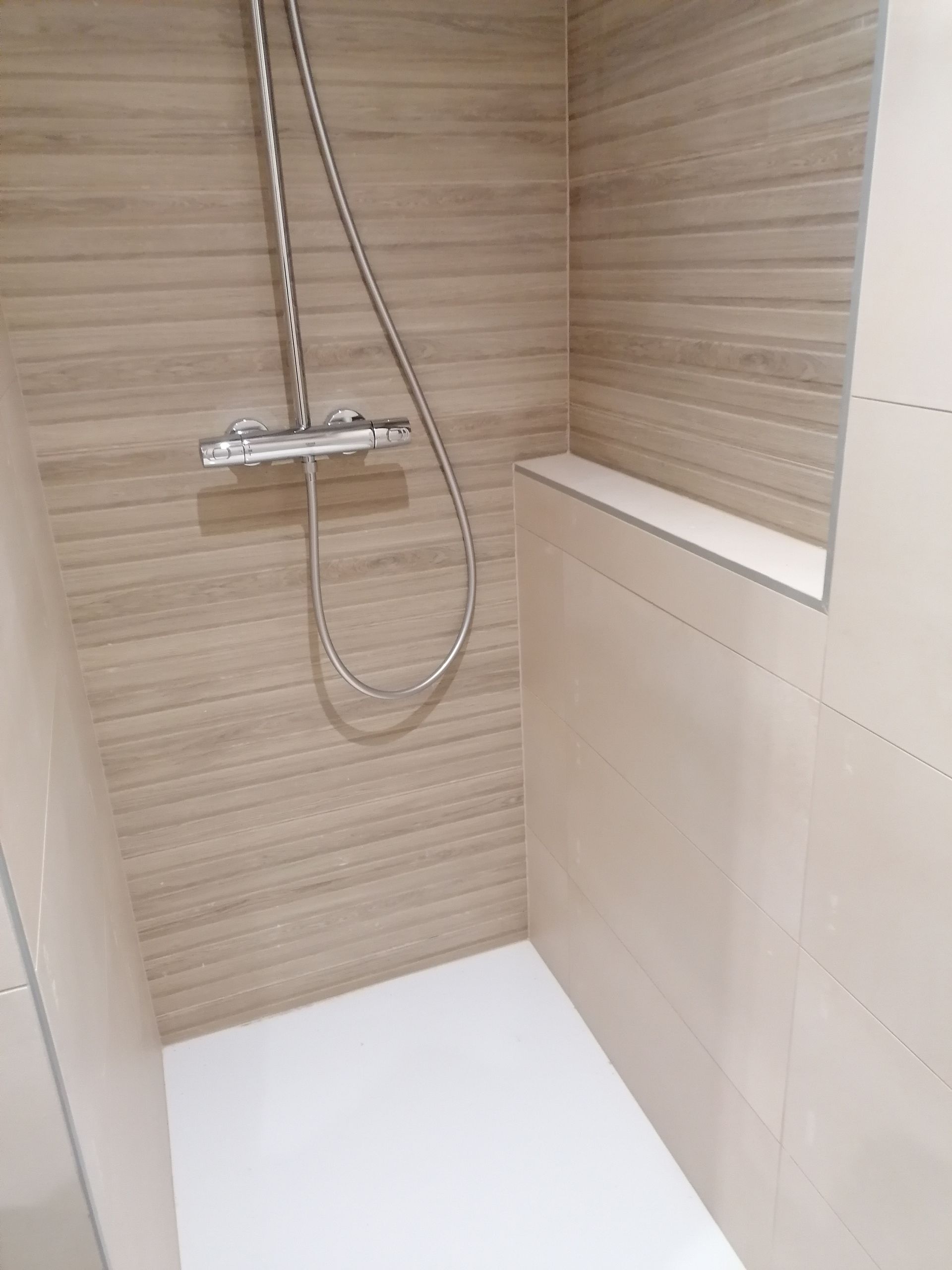 Une douche à l'italienne moderne avec des accents de carrelage clair imitation bois, une pomme de douche murale et un receveur carrelé blanc.
