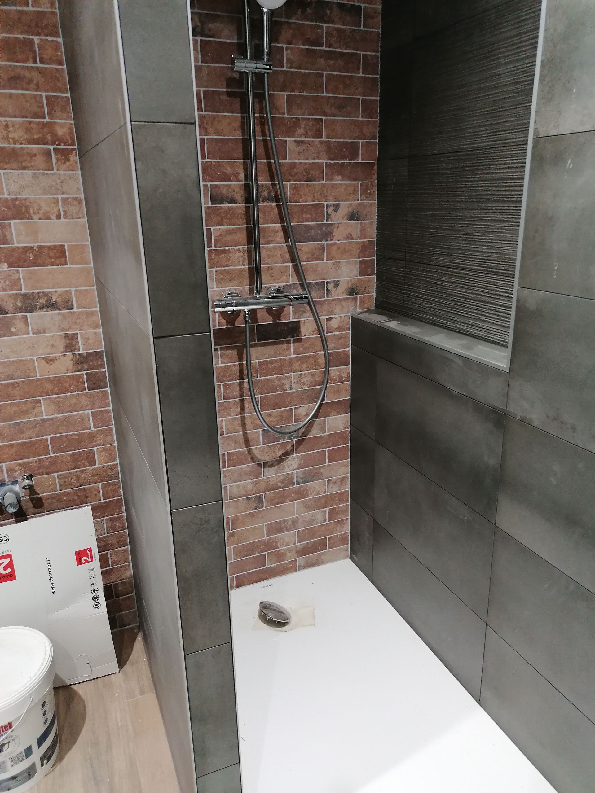 Cabine de douche avec receveur blanc, mur du fond à motif de briques, parois latérales carrelées gris foncé et pommeau de douche effet pluie apparent.