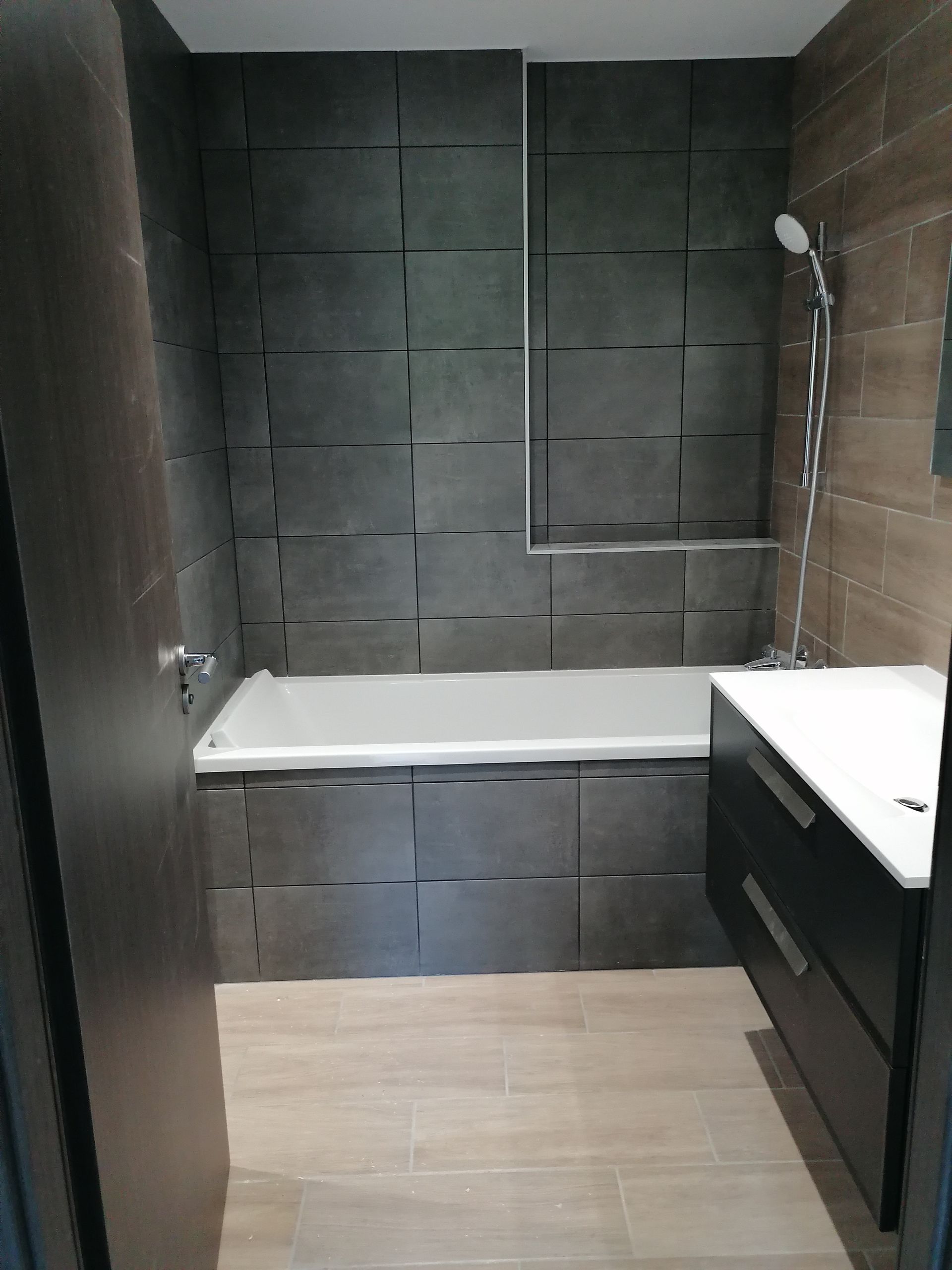 Une salle de bains moderne avec des murs carrelés gris foncé, une baignoire blanche, un sol carrelé clair et un meuble-lavabo noir.