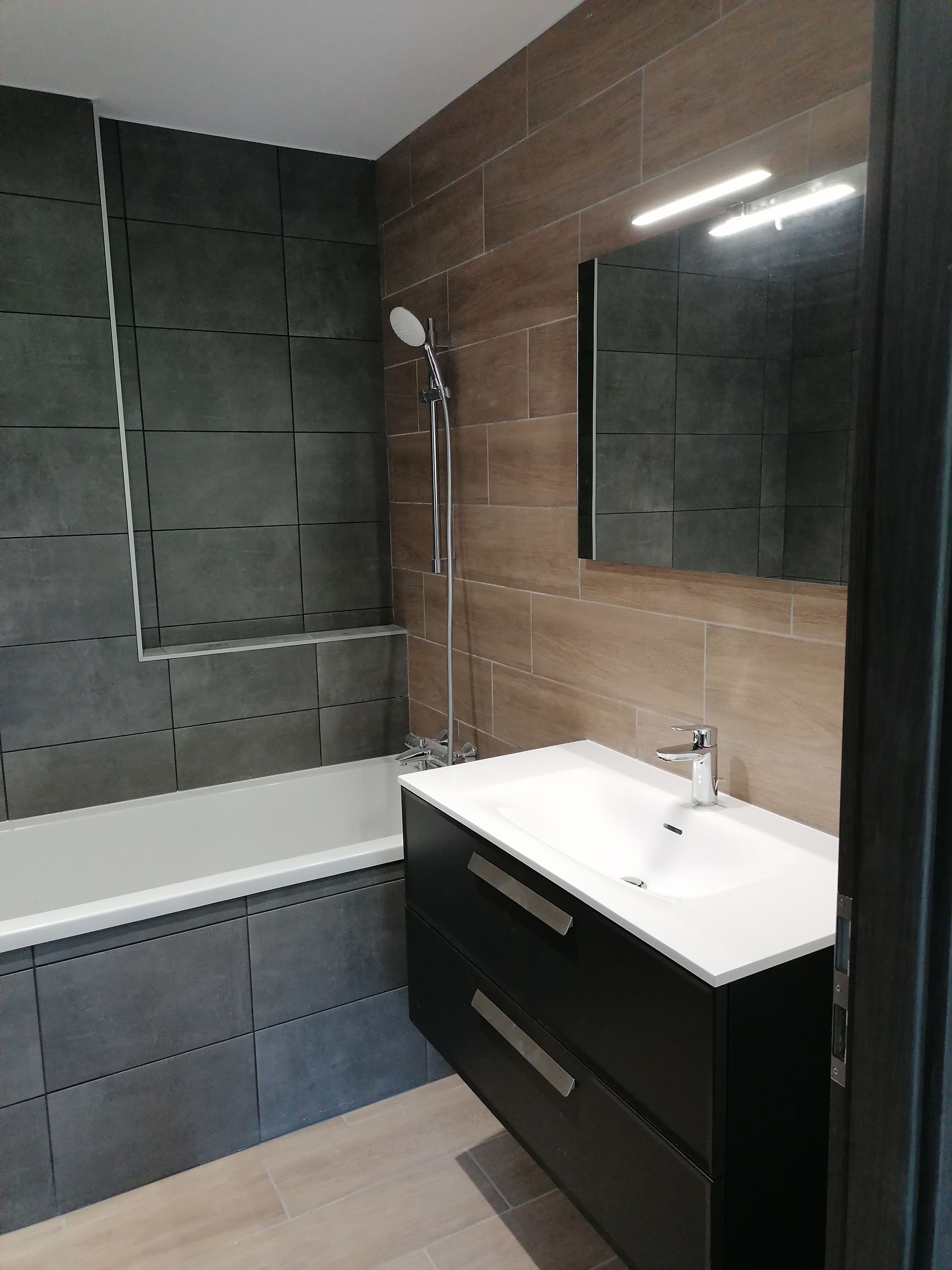 Une salle de bains moderne avec un meuble-lavabo suspendu foncé, un lavabo blanc, une baignoire carrelée de gris et des carreaux muraux imitation bois avec un miroir.