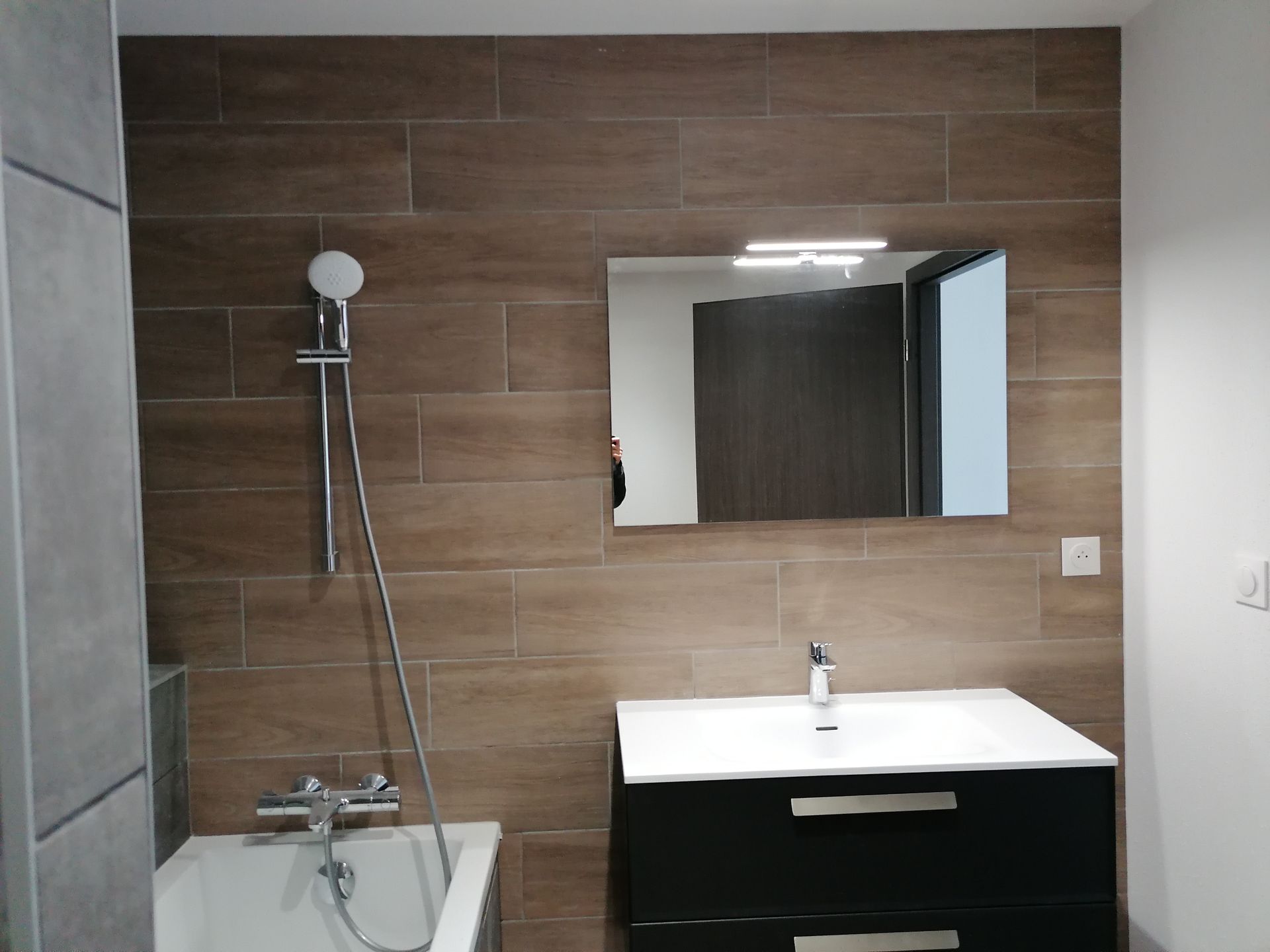 Salle de bains avec mur carrelé imitation bois, meuble-lavabo noir avec lavabo blanc, miroir éclairé et robinetterie de douche chromée.