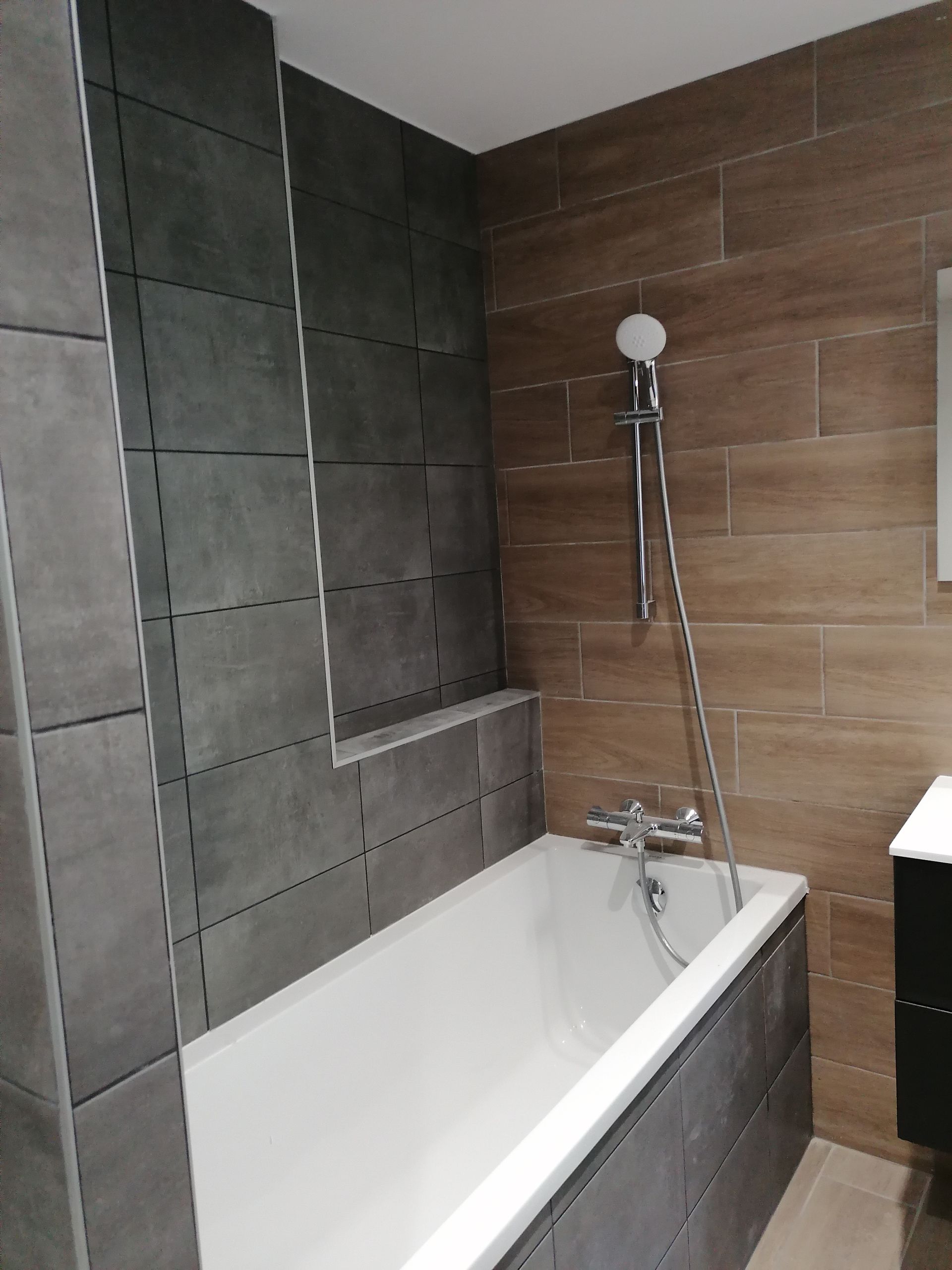 Une baignoire blanche dans une salle de bains moderne avec des murs carrelés gris autour de la baignoire et des carreaux muraux imitation bois derrière la pomme de douche.