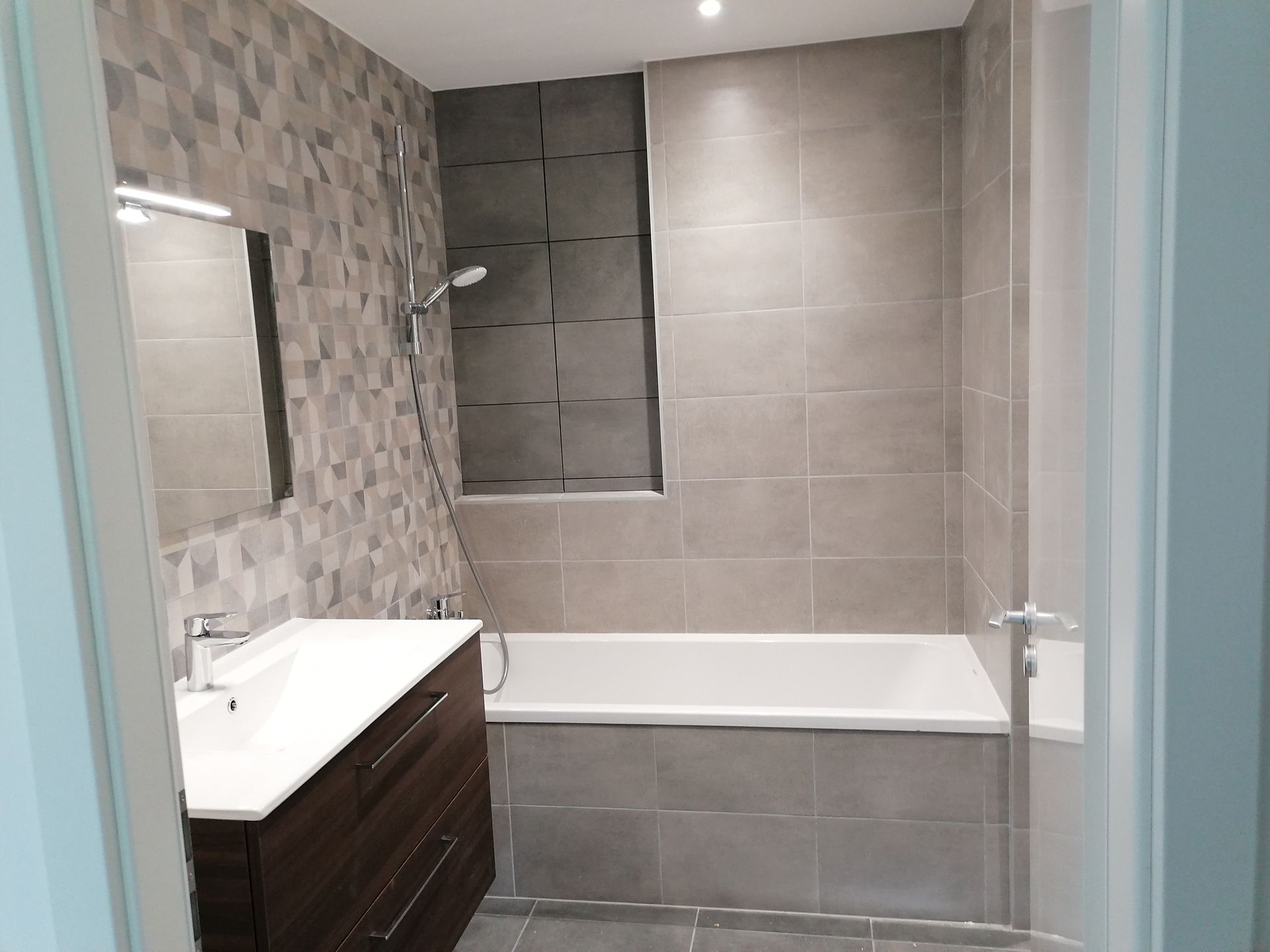 Salle de bains moderne avec meuble-lavabo en bois foncé, lavabo blanc, baignoire et murs carrelés gris.