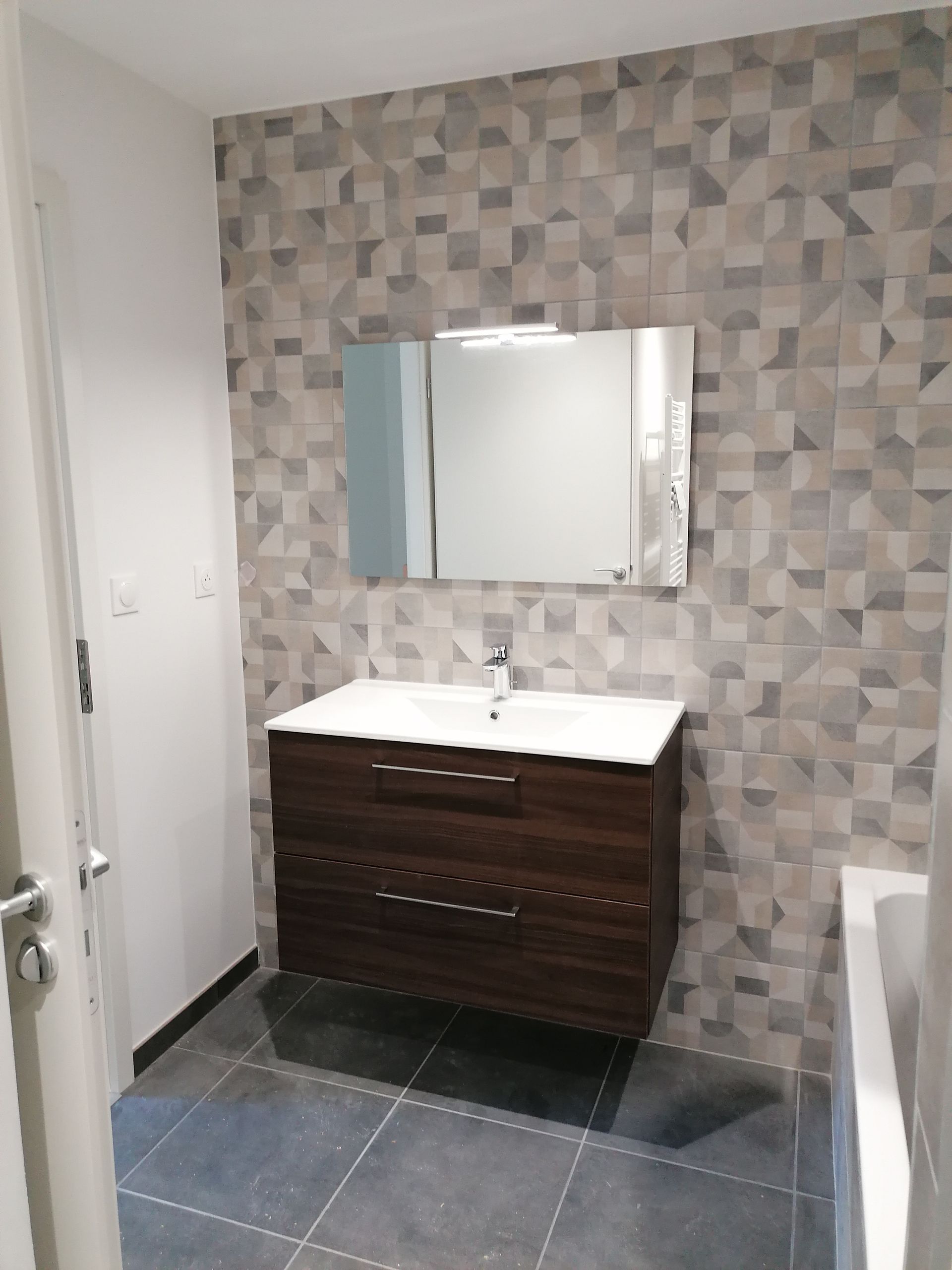 Salle de bains moderne avec meuble-lavabo suspendu en bois foncé, lavabo blanc, miroir rectangulaire et carrelage mural géométrique gris texturé.