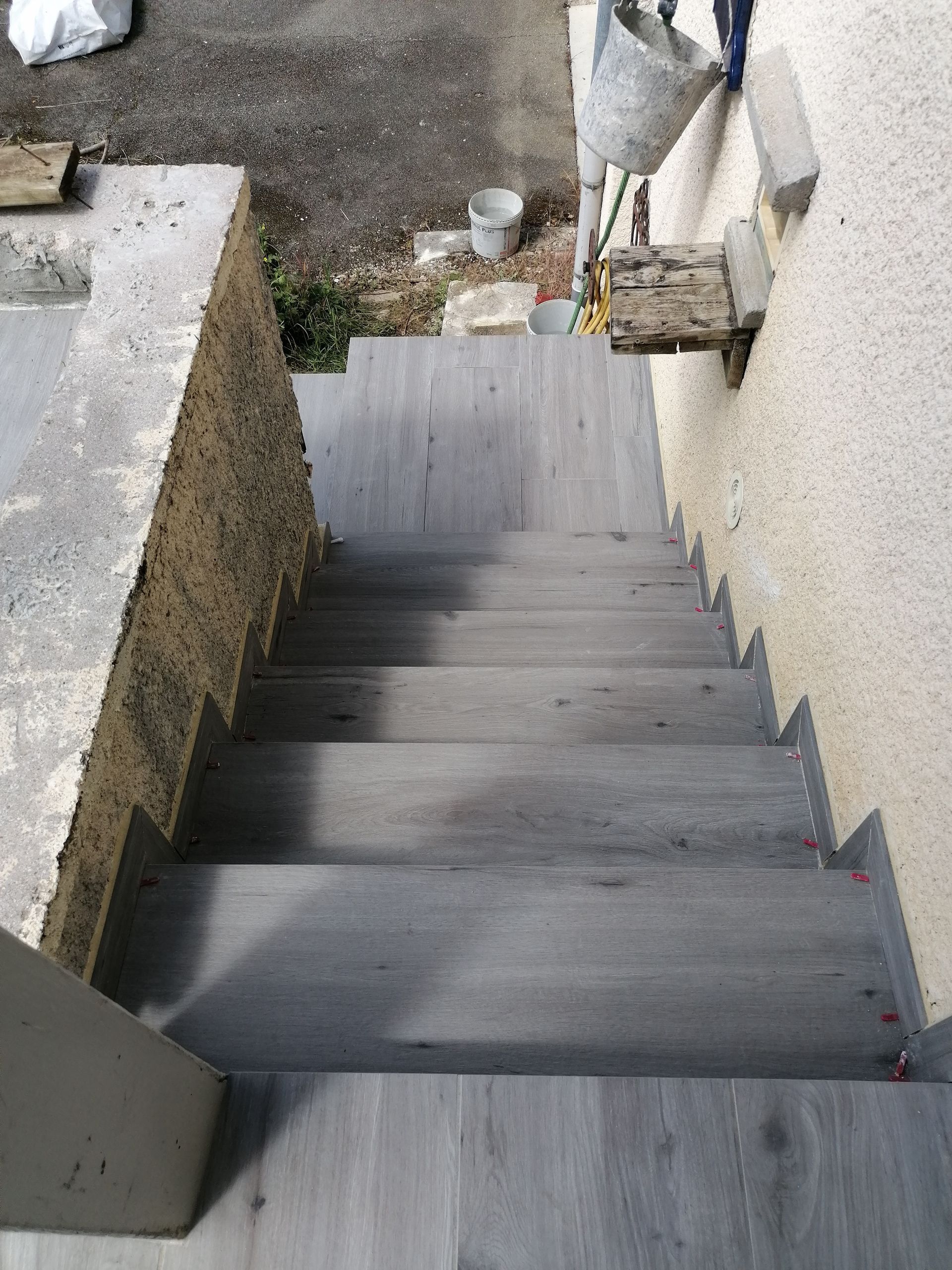 Vue plongeante d'un escalier recouvert de carrelage gris imitation bois, situé entre deux murs.