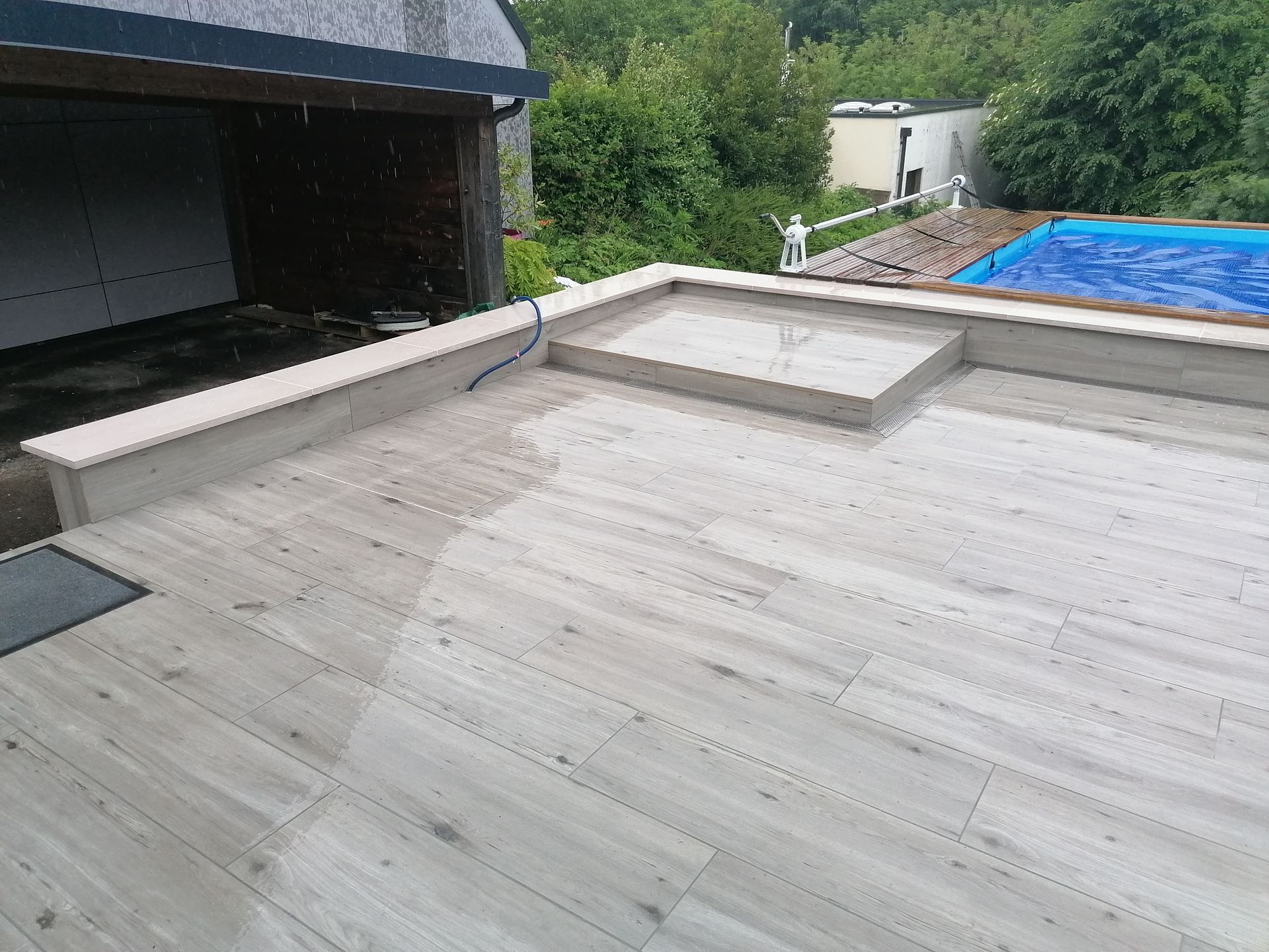 Une terrasse extérieure spacieuse avec des carreaux clairs imitation bois, un banc en pierre intégré et une piscine attenante.