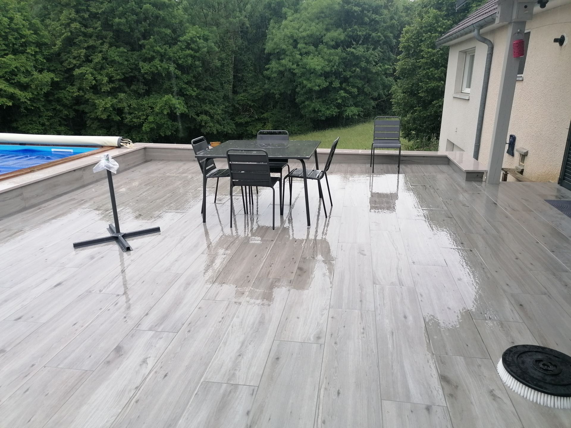 Terrasse ombragée avec table et chaises. Piscine, ciel couvert, carrelage gris clair imitation bois.