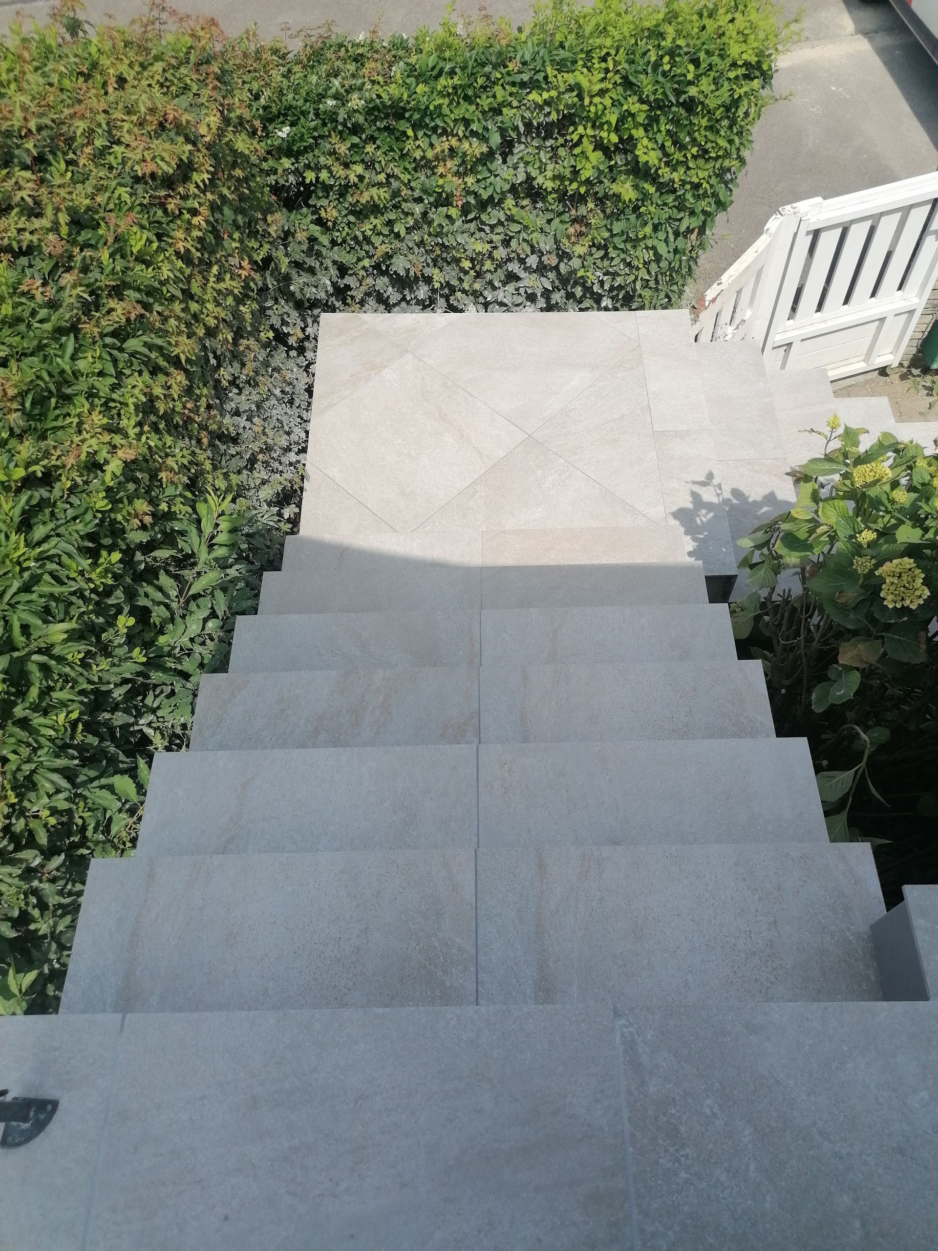 Vue en plongée d'un escalier en pierre gris clair descendant vers un chemin pavé, encadré par des buissons verts et une clôture blanche.