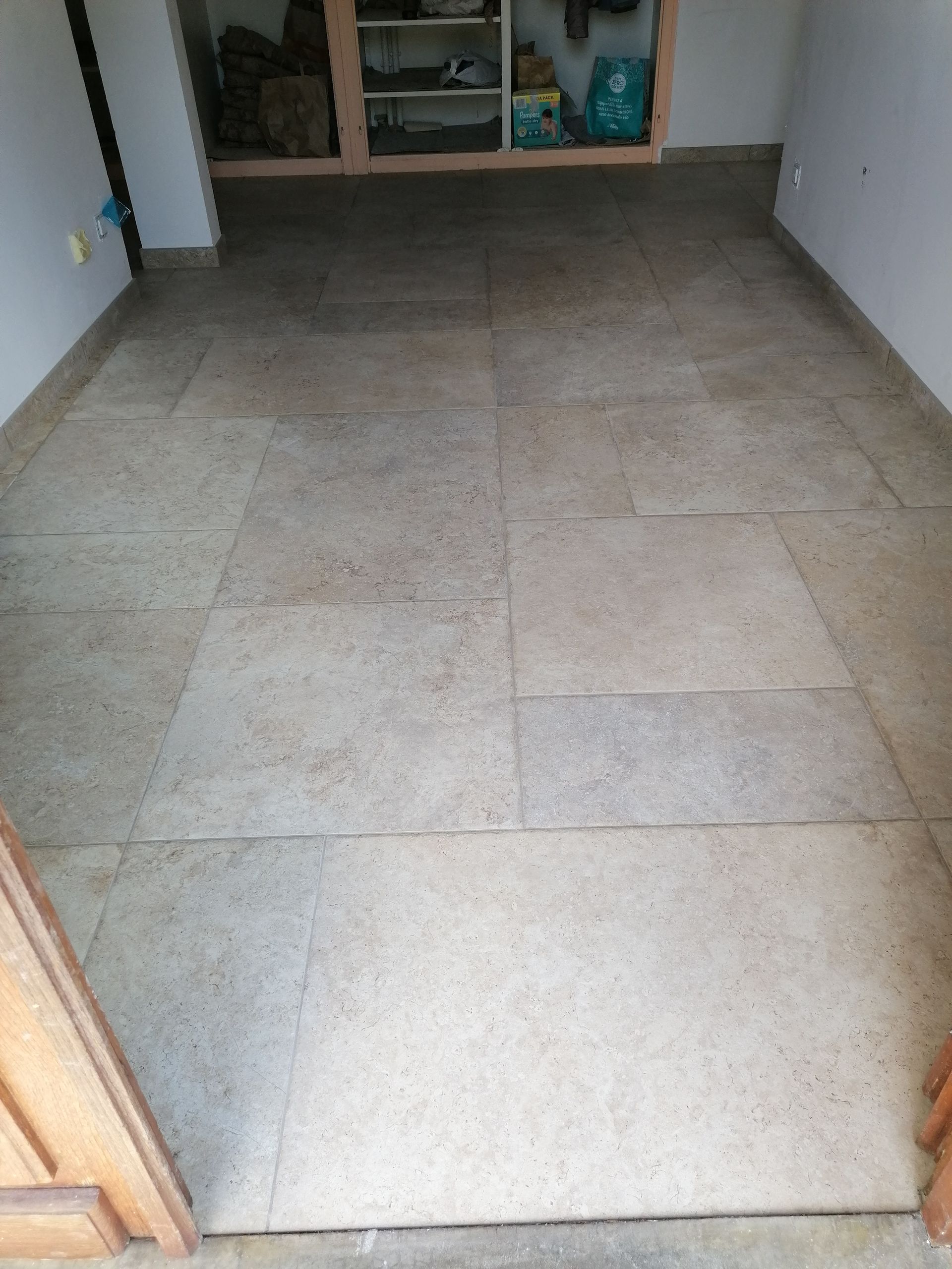 Sol carrelé beige clair dans une pièce. Un cadre de porte en bois est visible au premier plan. Des étagères se devinent à l'arrière-plan.