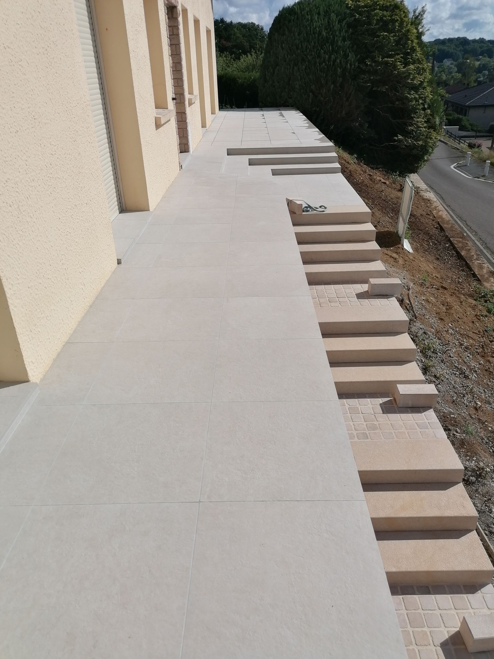 Terrasse en pierre beige avec des marches descendant vers une zone en pente et la rue.