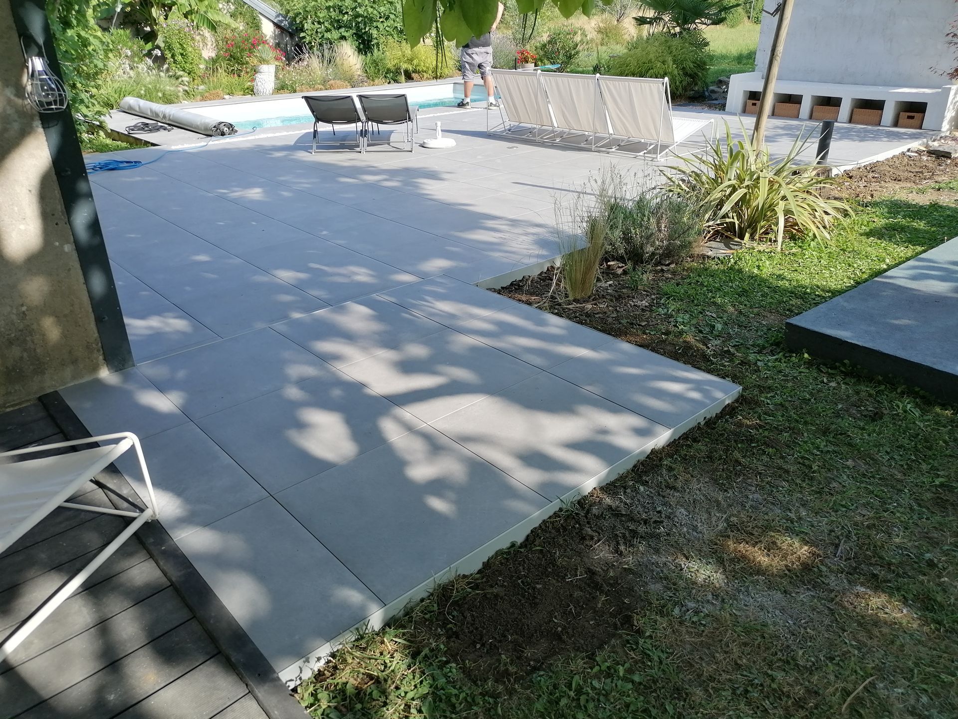 Une terrasse gris clair avec du mobilier de jardin jouxtant une piscine et un jardin par une journée ensoleillée.