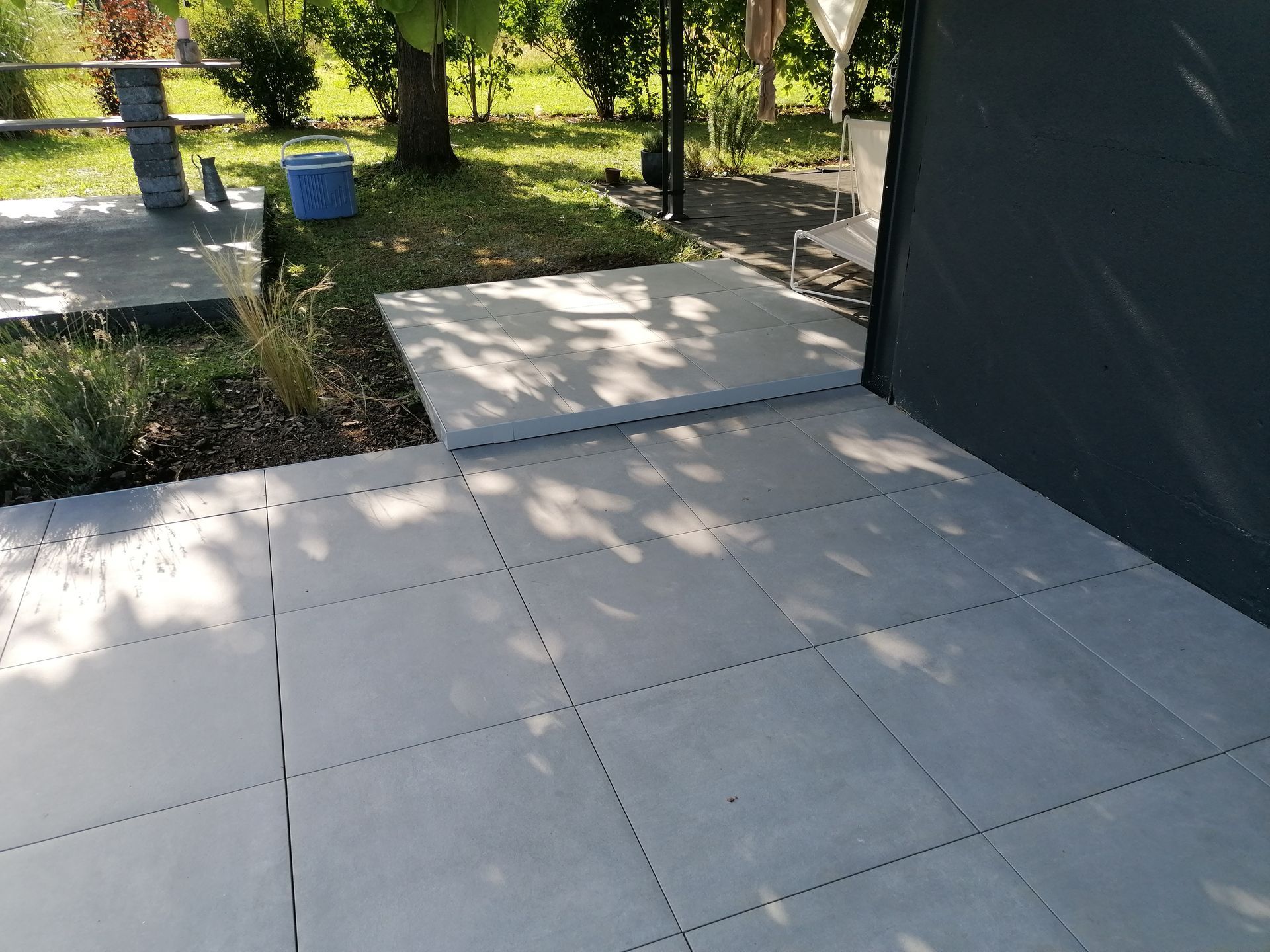 Terrasse carrelée de gris, baignée d'une lumière tamisée, donnant sur une pelouse verdoyante et offrant une vue partielle sur une structure.