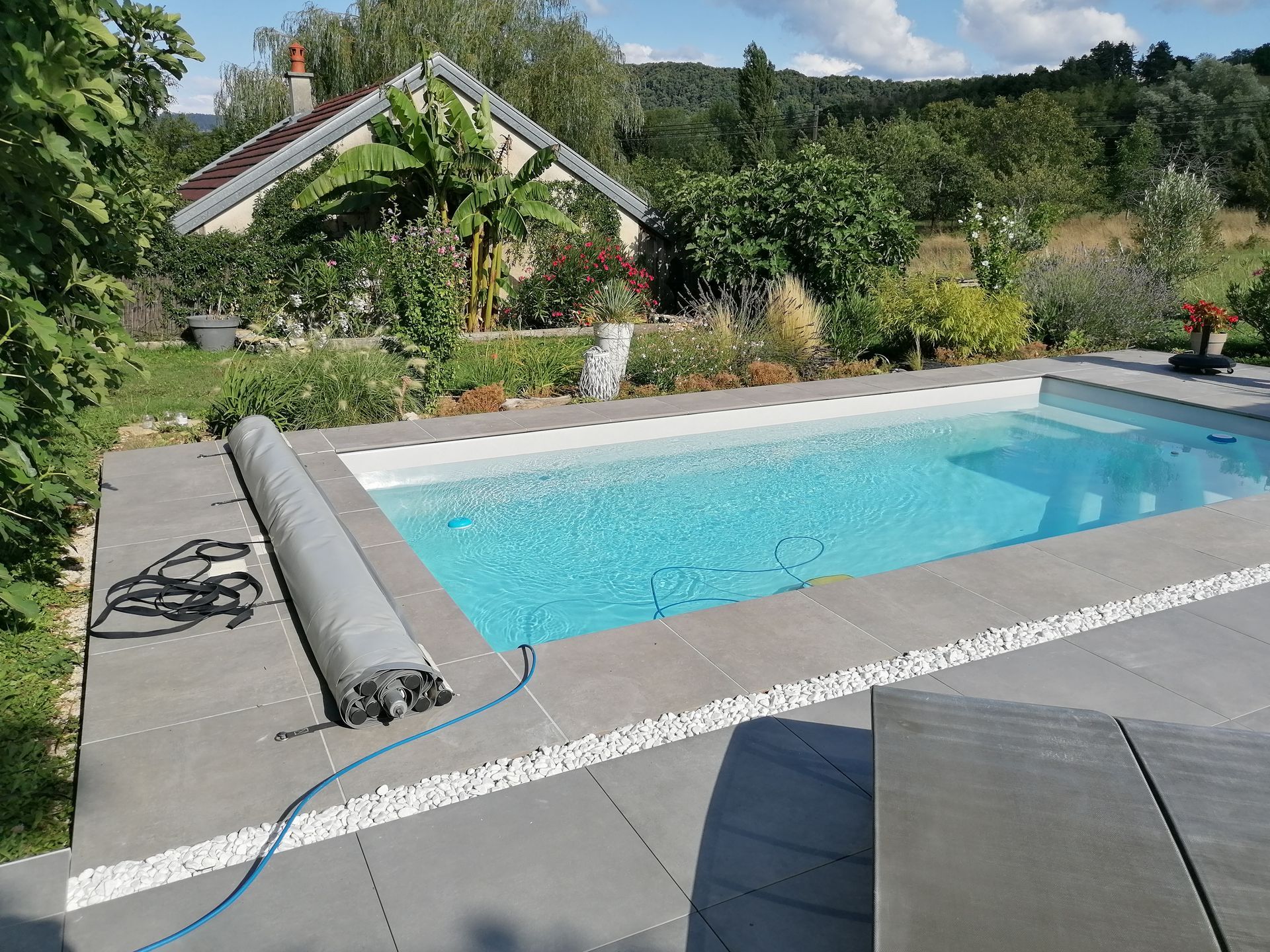 Piscine rectangulaire à l'eau bleue et terrasse en pierre grise, entourée de verdure et d'une maison.