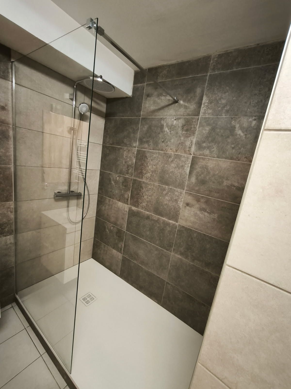 Une douche à l'italienne moderne avec des murs carrelés à la texture foncée et une paroi en verre, équipée d'une pomme de douche effet pluie et d'une bonde.