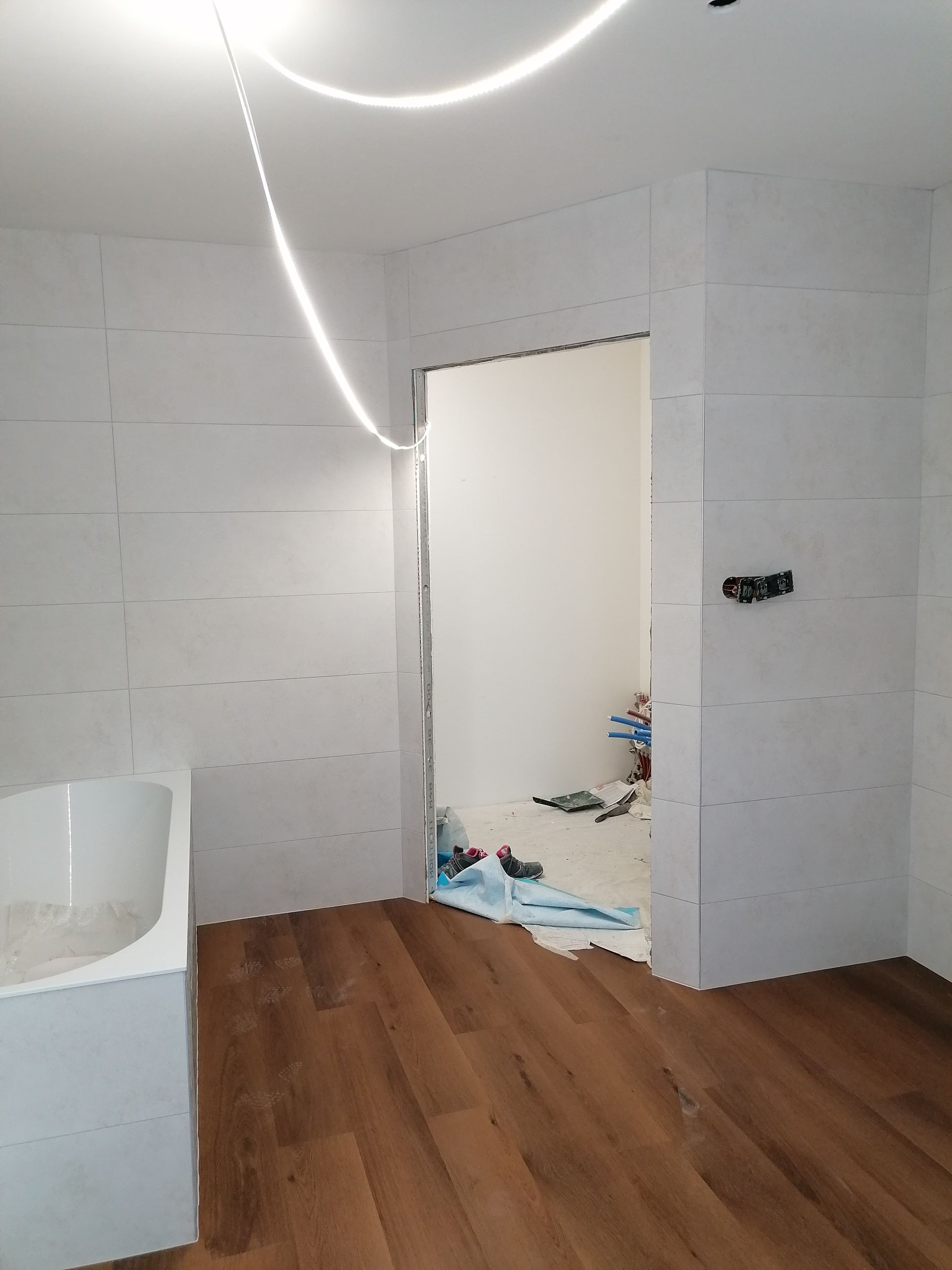 Une salle de bains en rénovation avec des murs carrelés blancs, un sol imitation bois, une baignoire blanche et une porte ouverte donnant sur une autre pièce.