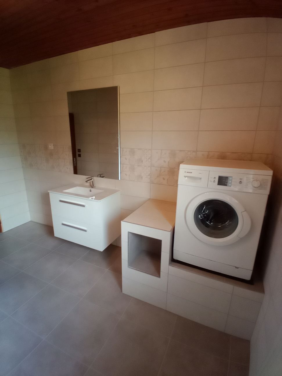Salle de bains avec meuble-lavabo blanc, miroir, machine à laver et carrelage gris au sol et aux murs.
