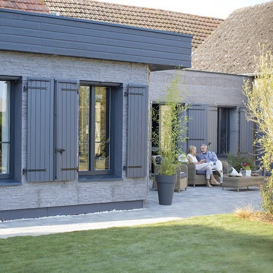 Un couple assis dans un jardin devant une maison en pierres avec des volets battants de la marque Somfy