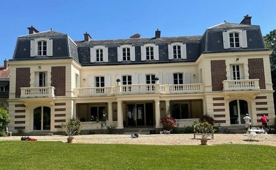 Élégant manoir à deux étages avec un long balcon, des murs beiges et un toit marron sur fond de ciel bleu.