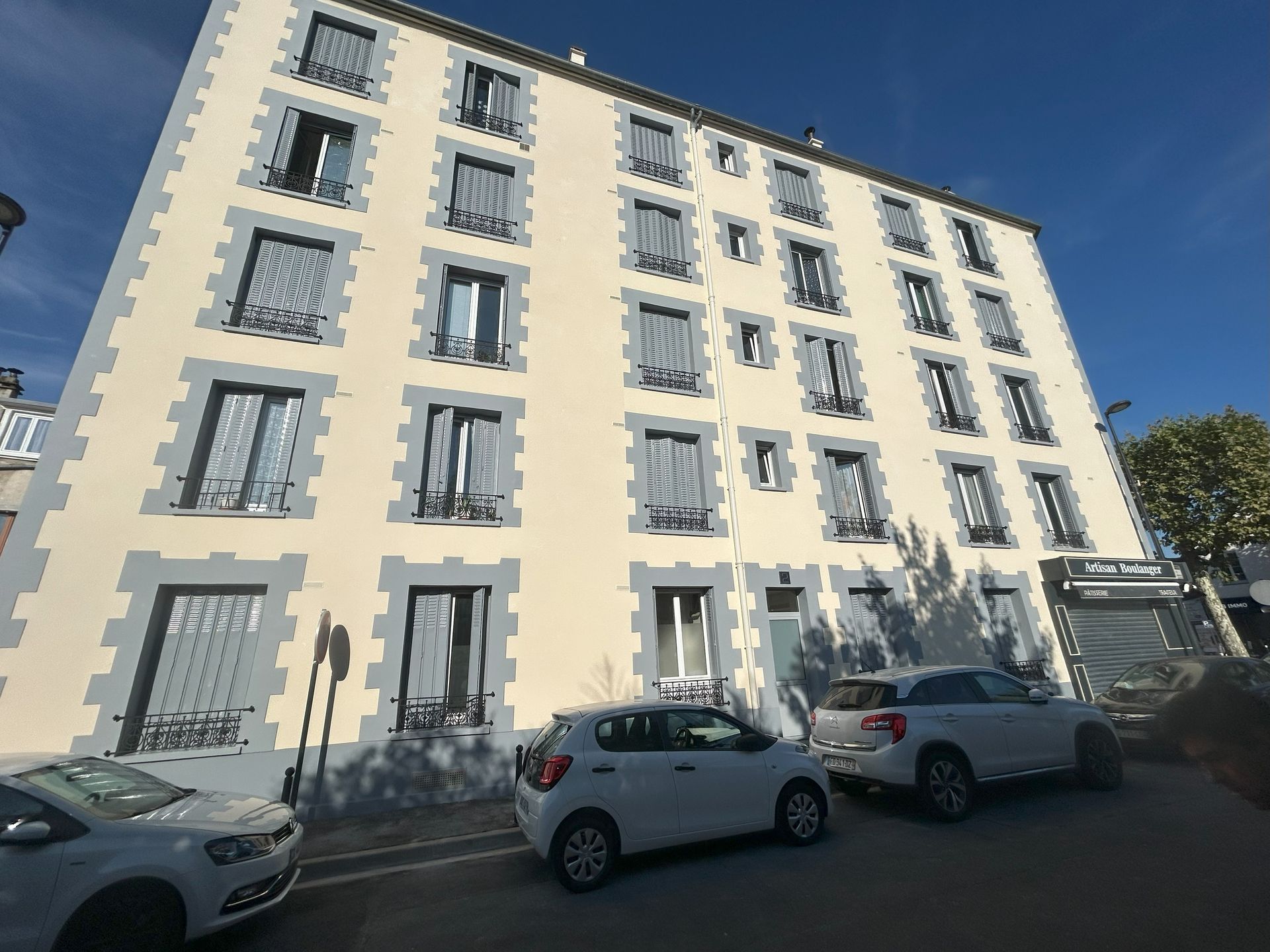 Un immeuble d'appartements à plusieurs étages avec des volets bleus et des voitures garées par une journée ensoleillée.