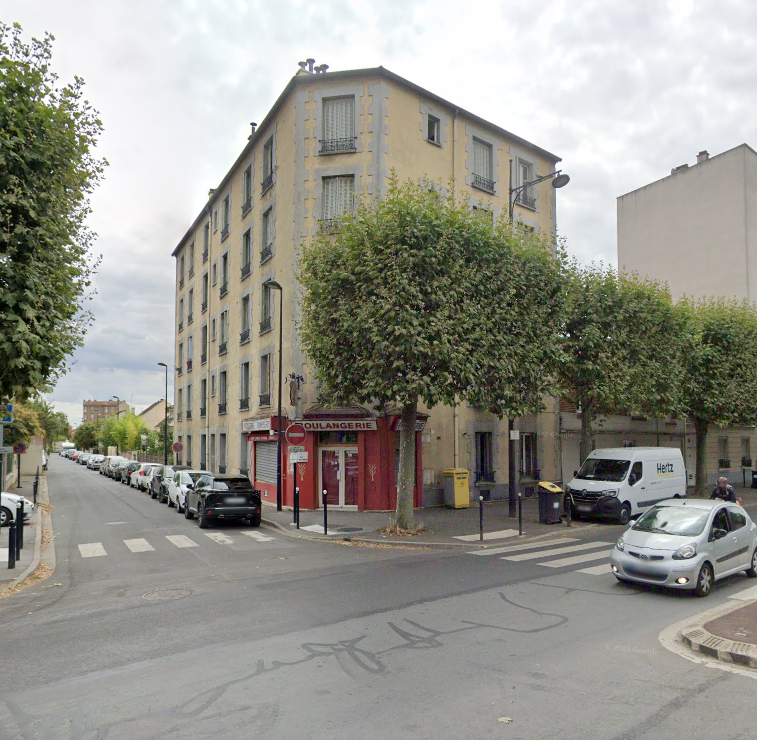 Coin de rue avec un bâtiment beige. Voitures garées dans la rue. Arbres et boutique aux touches de rouge.