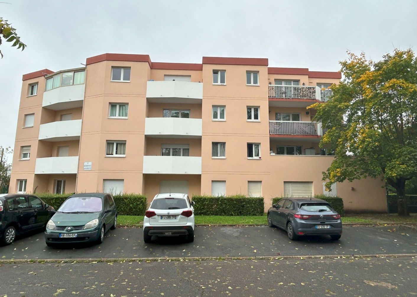 Immeuble d'habitation, façade rose clair avec balcons. Voitures garées devant. Temps couvert.