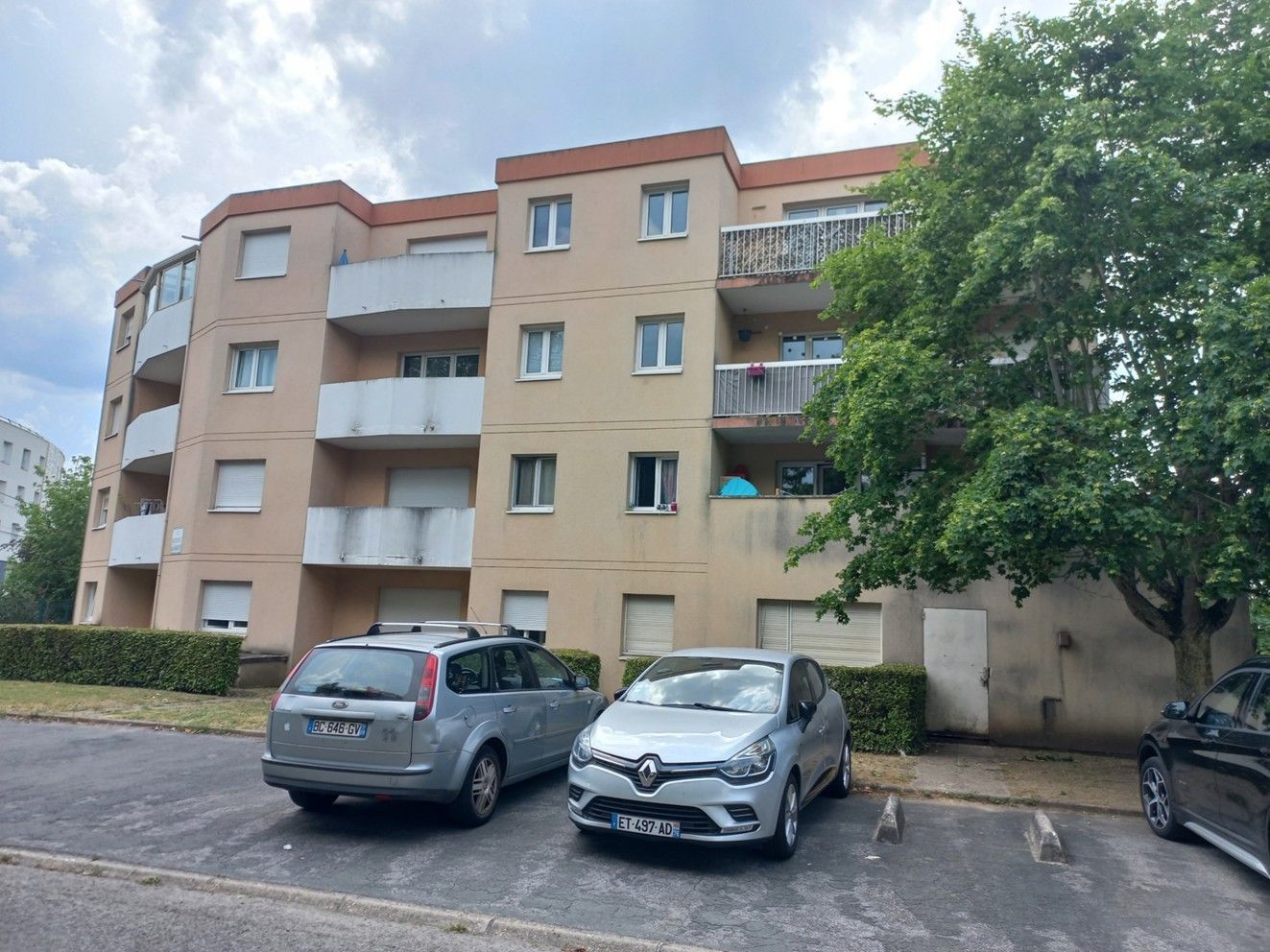 Immeuble résidentiel avec voitures garées devant. Façade beige, balcons et arbre à droite.