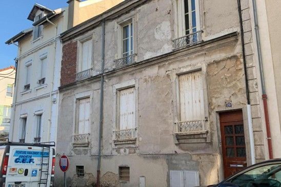 Bâtiment de deux étages avec stuc écaillé, volets blancs et porte marron.