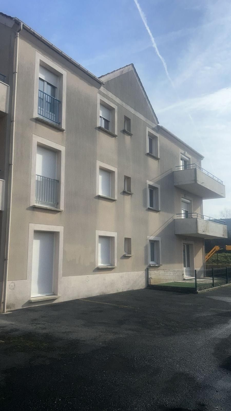 Immeuble de trois étages à la façade beige, aux fenêtres et portes blanches et à un petit balcon. Au premier plan, asphalte et ciel dégagé.