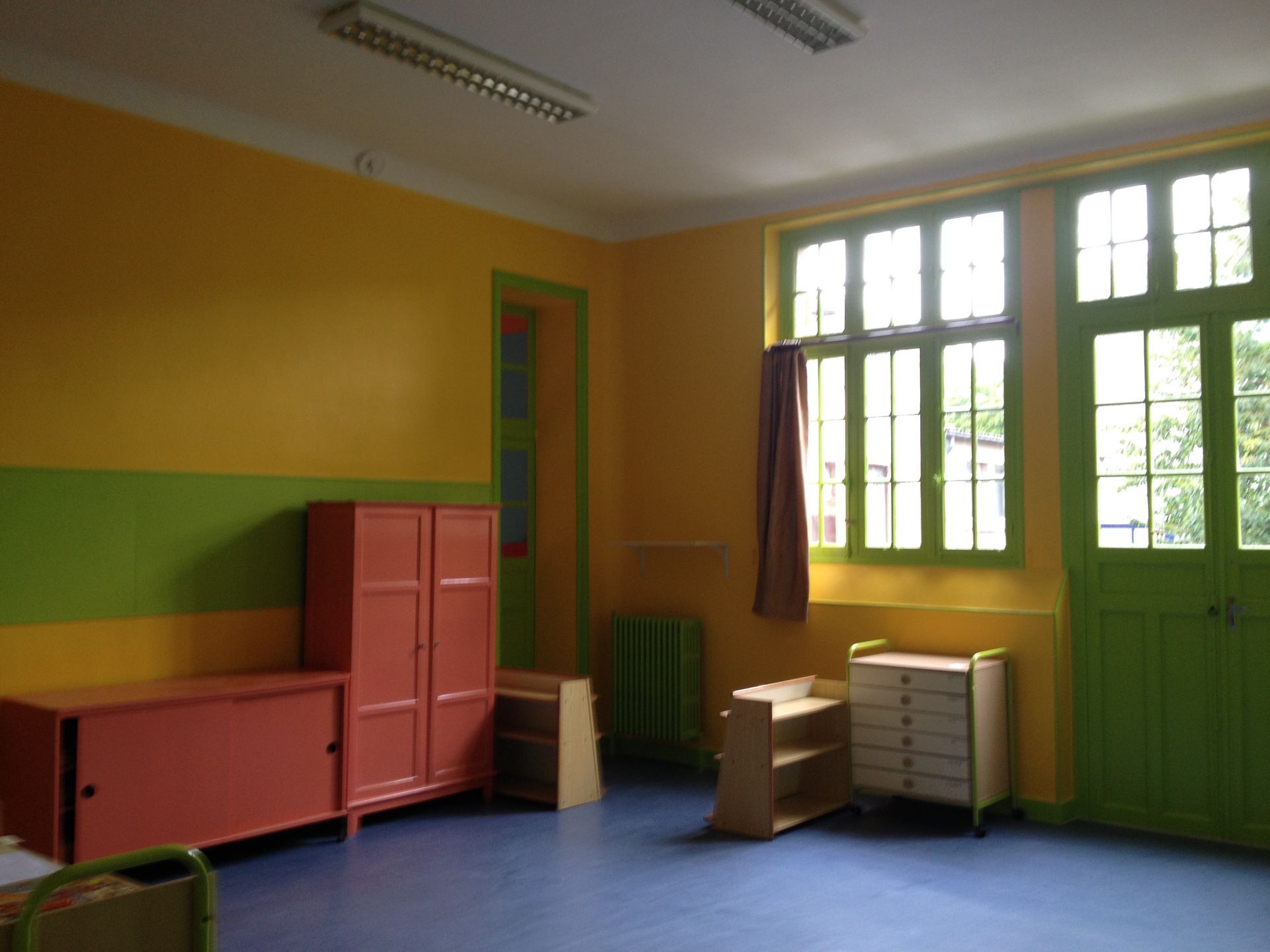 Salle de classe vide avec des murs jaunes et verts, un sol bleu, des armoires colorées et une grande fenêtre.