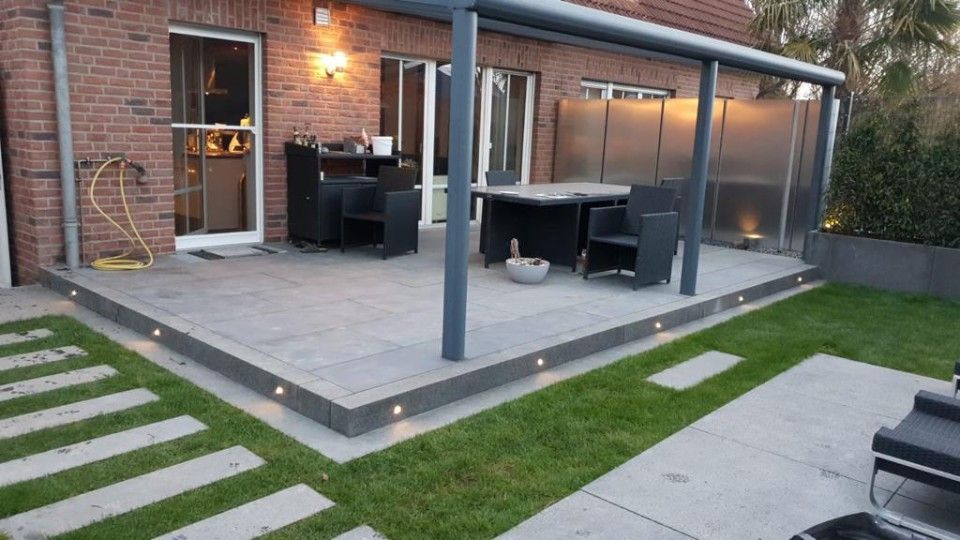 Eine Terrasse mit Tisch und Stühlen unter einem Vordach vor einem Backsteinhaus.