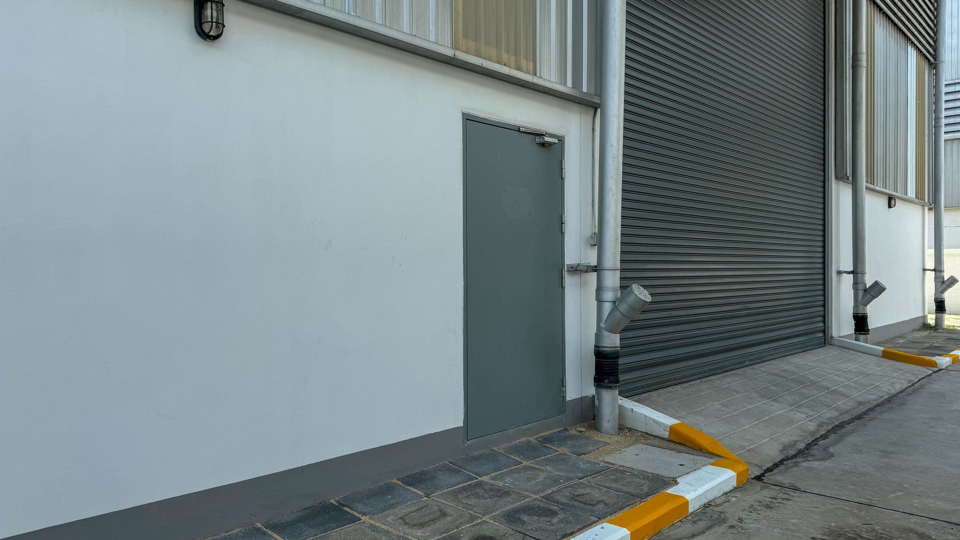 Vue latérale du mur gris d'un bâtiment avec une porte métallique et une rampe à côté d'une entrée fermée par un portail.