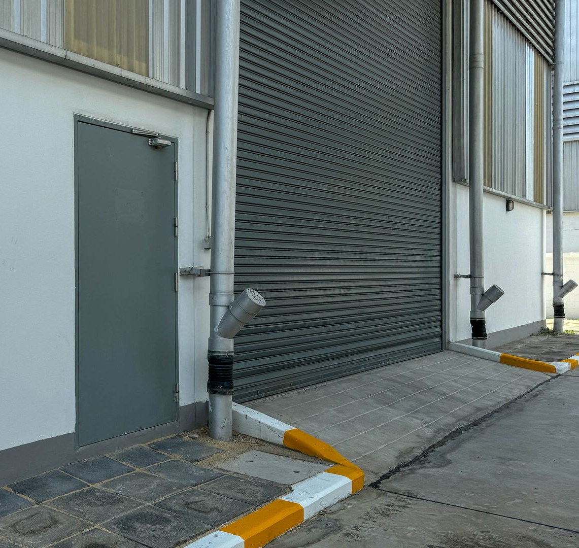 Entrée latérale d'un bâtiment industriel avec porte grise, rideau métallique et rampes d'accès le long du trottoir.