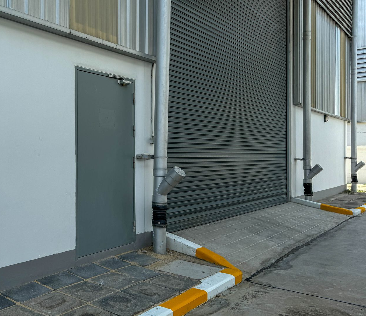 Extérieur d'un bâtiment industriel avec porte grise, grand rideau métallique et marquages jaunes au sol le long d'un trottoir.